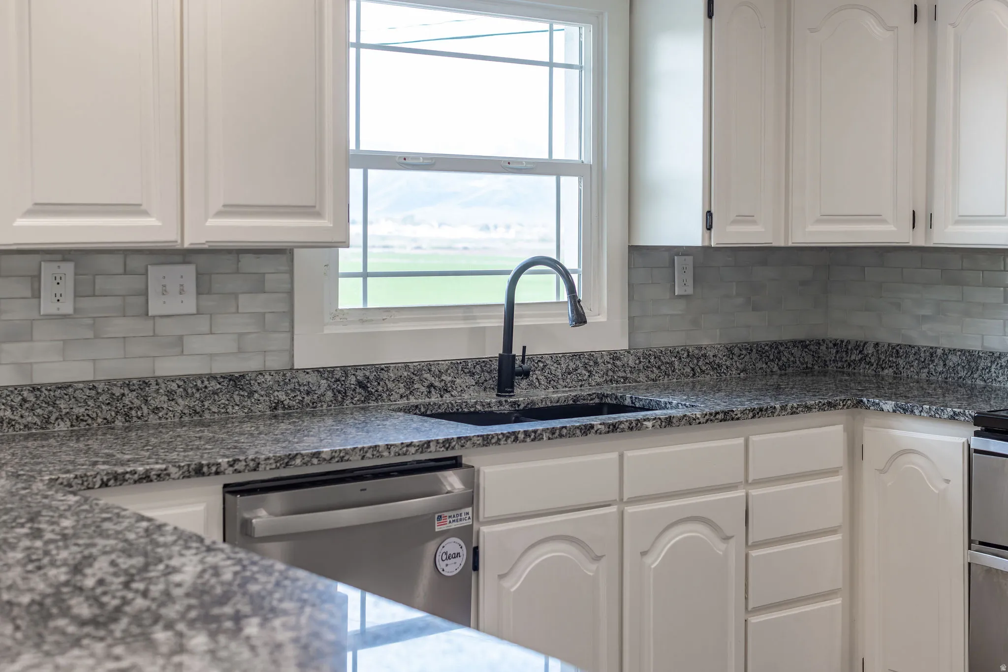 Kitchen with white cabinets, stainless steel dishwasher, dark stone countertops, backsplash, and plenty of natural light