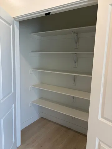 Interior closet featuring five white shelves, light-toned wood-finish flooring, and white trim work