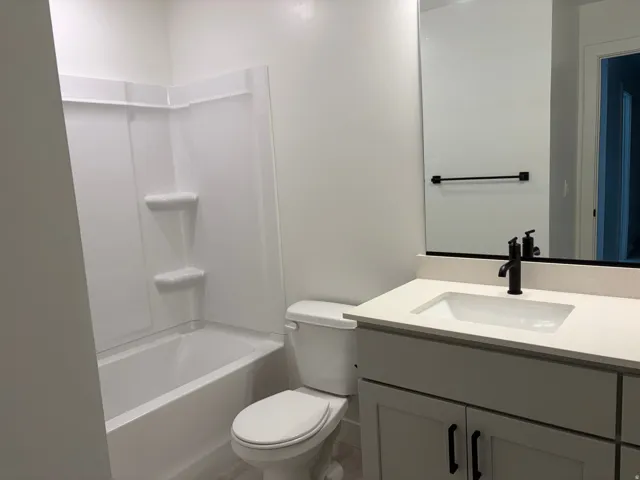 Bathroom featuring a white integrated tub and shower unit, a toilet, a vanity with a light-toned countertop, an undermount sink, and a black-finish faucet