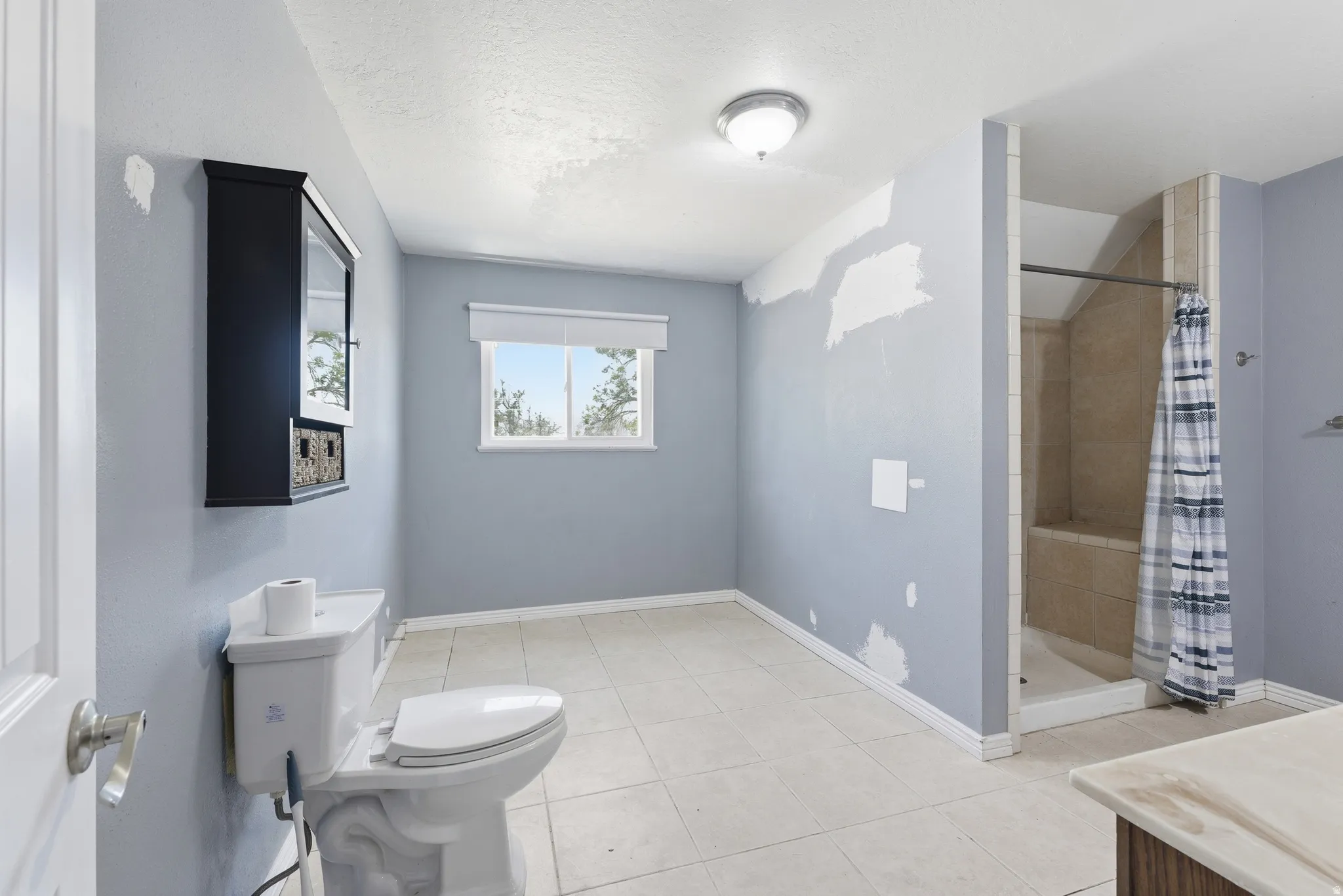 Full bathroom featuring a walk-in shower with tiled surround and built-in bench, a window with a roller shade, tile flooring, and a wall-mounted storage cabinet