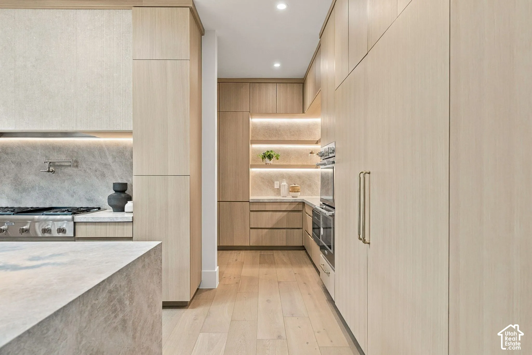 Kitchen featuring flat-panel light wood cabinetry, stone-finish countertops and backsplash, integrated double wall ovens, wood-finish flooring, and under-cabinet recessed lighting