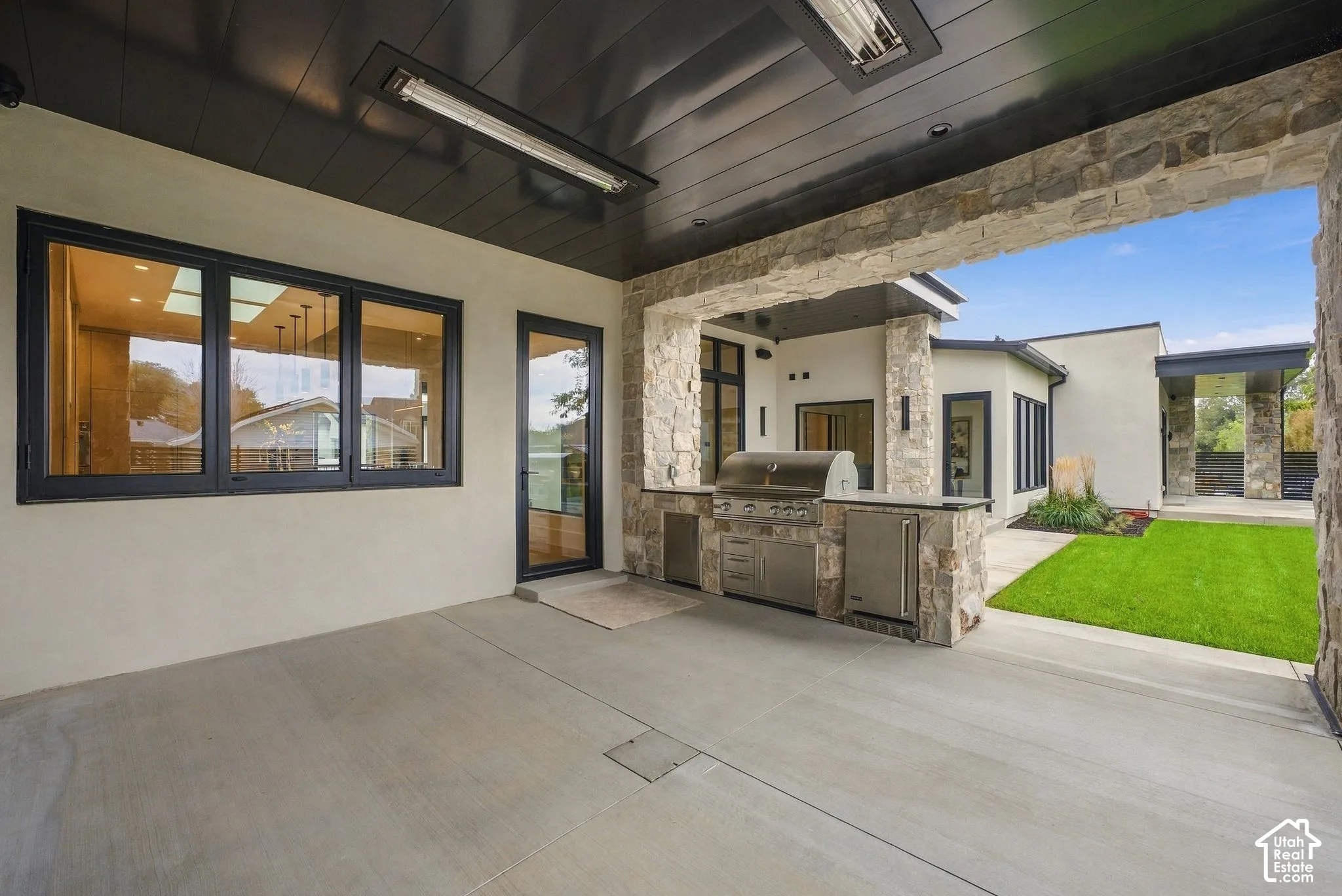 Covered outdoor patio featuring a built-in stainless steel grill station with stone masonry, dark metal ceiling panels, integrated overhead heaters, and black-framed exterior windows and doors