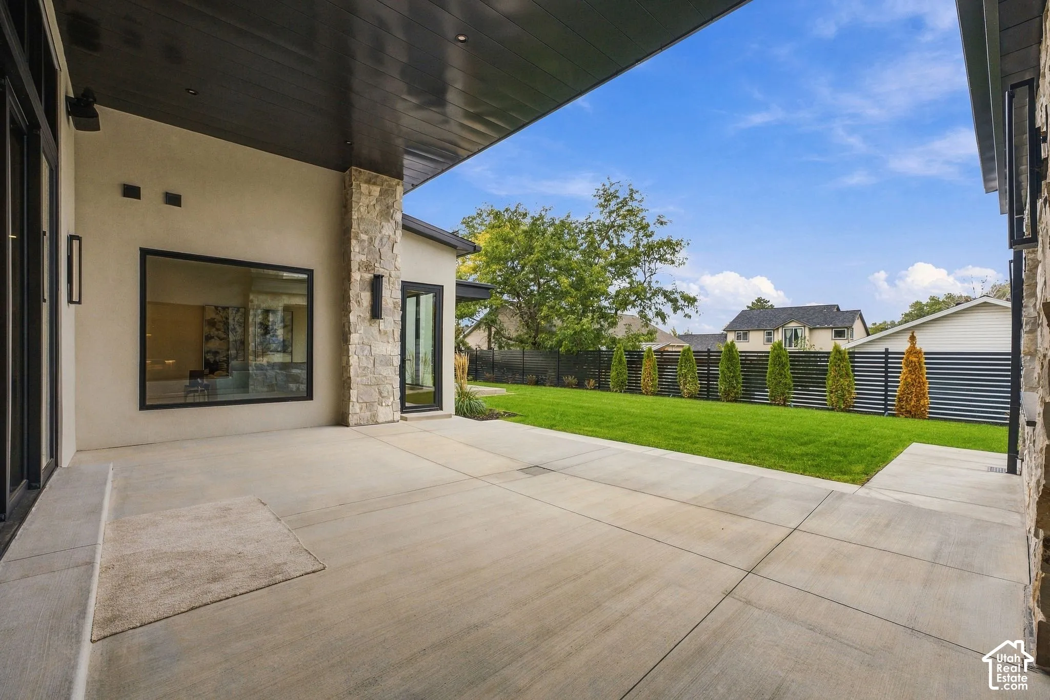 Covered concrete patio featuring a dark-finished ceiling, natural stone column accents, and a large picture window, opening to a fenced backyard with a lawn