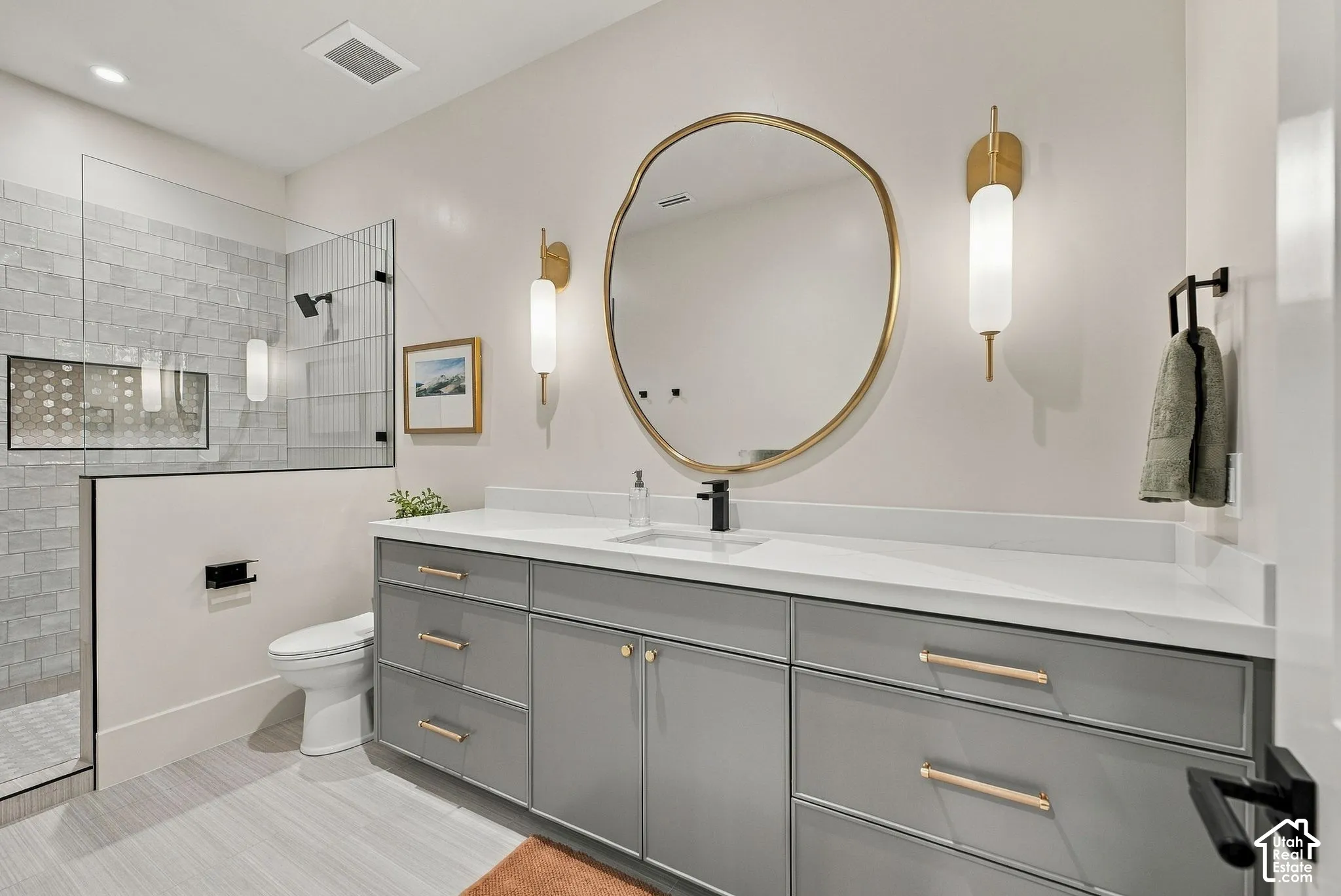 Dual-sink vanity with matte grey cabinetry, light-toned stone countertop, organic-shaped gold-rimmed mirror, paired glass-and-gold wall sconces, and walk-in shower with stone tile surround