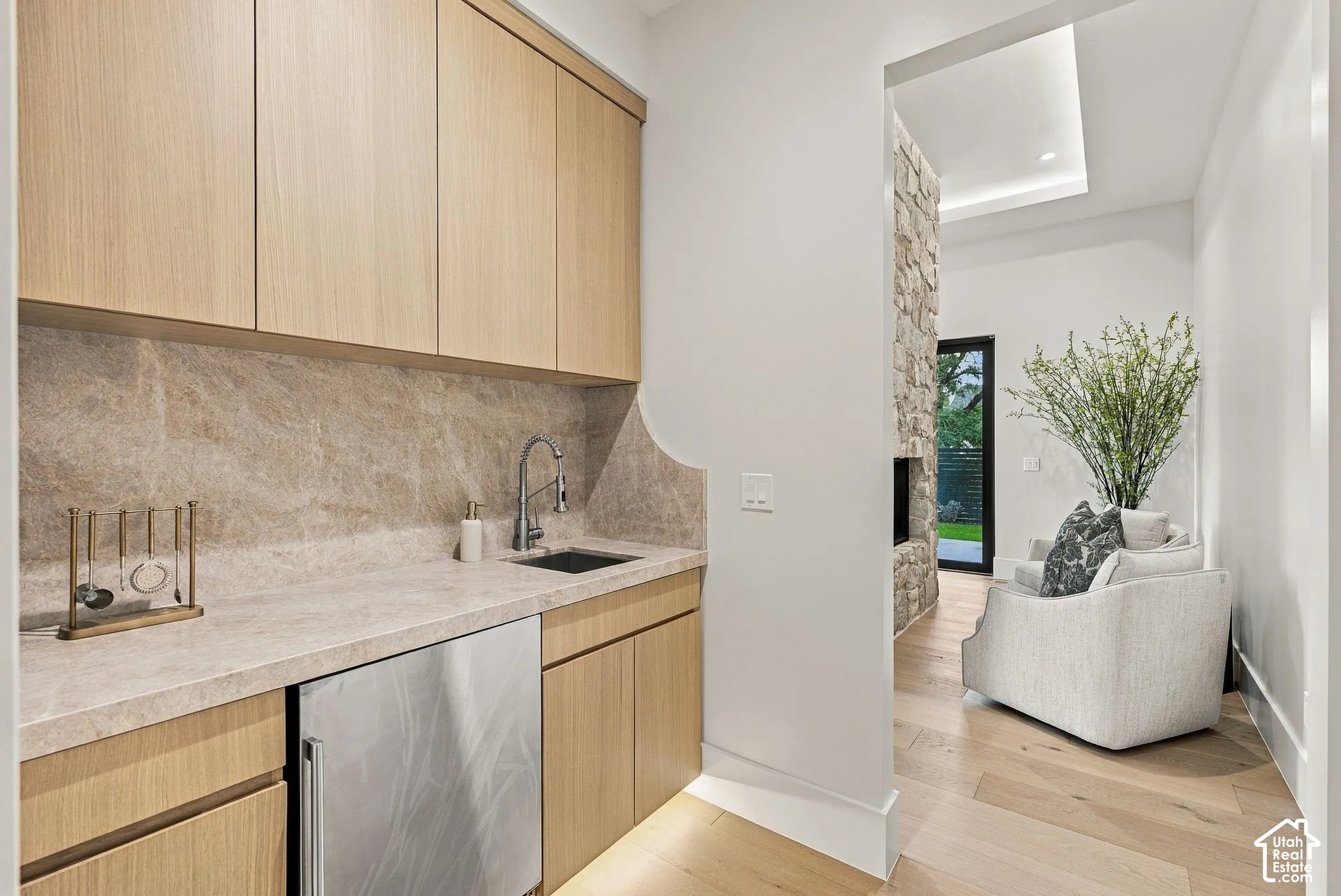 Wet bar featuring flat-panel wood cabinetry, natural stone backsplash and countertop, stainless steel under-mount sink with gooseneck faucet, and built-in stainless steel under-counter refrigerator