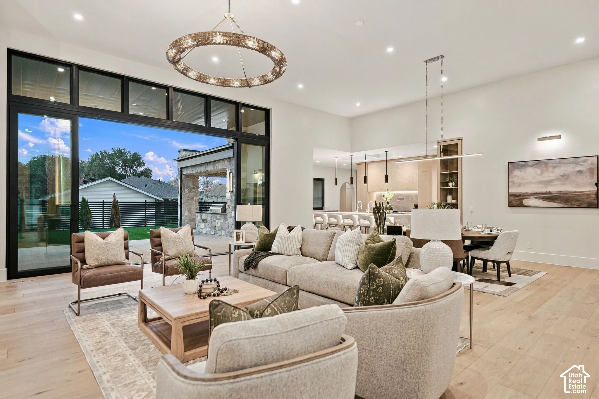 Open-concept interior featuring large-format multi-slide glass doors to allow indoor/ outdoor living, light-toned wood-finish flooring, recessed lighting, and a prominent circular statement chandelier
