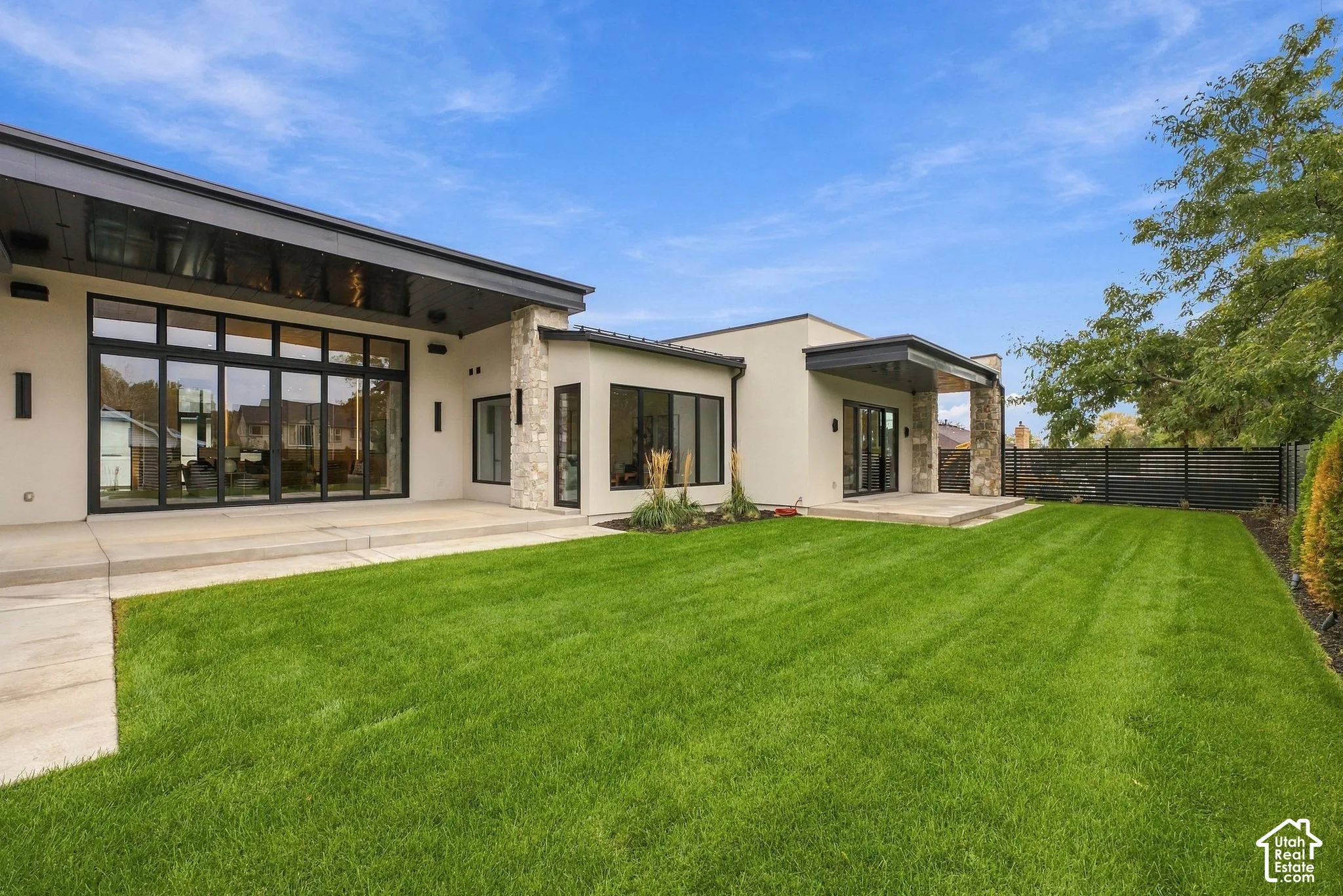 Modern exterior featuring smooth stucco siding, stone masonry accents, expansive floor-to-ceiling multi-panel glass doors, covered concrete patio, and a manicured lawn