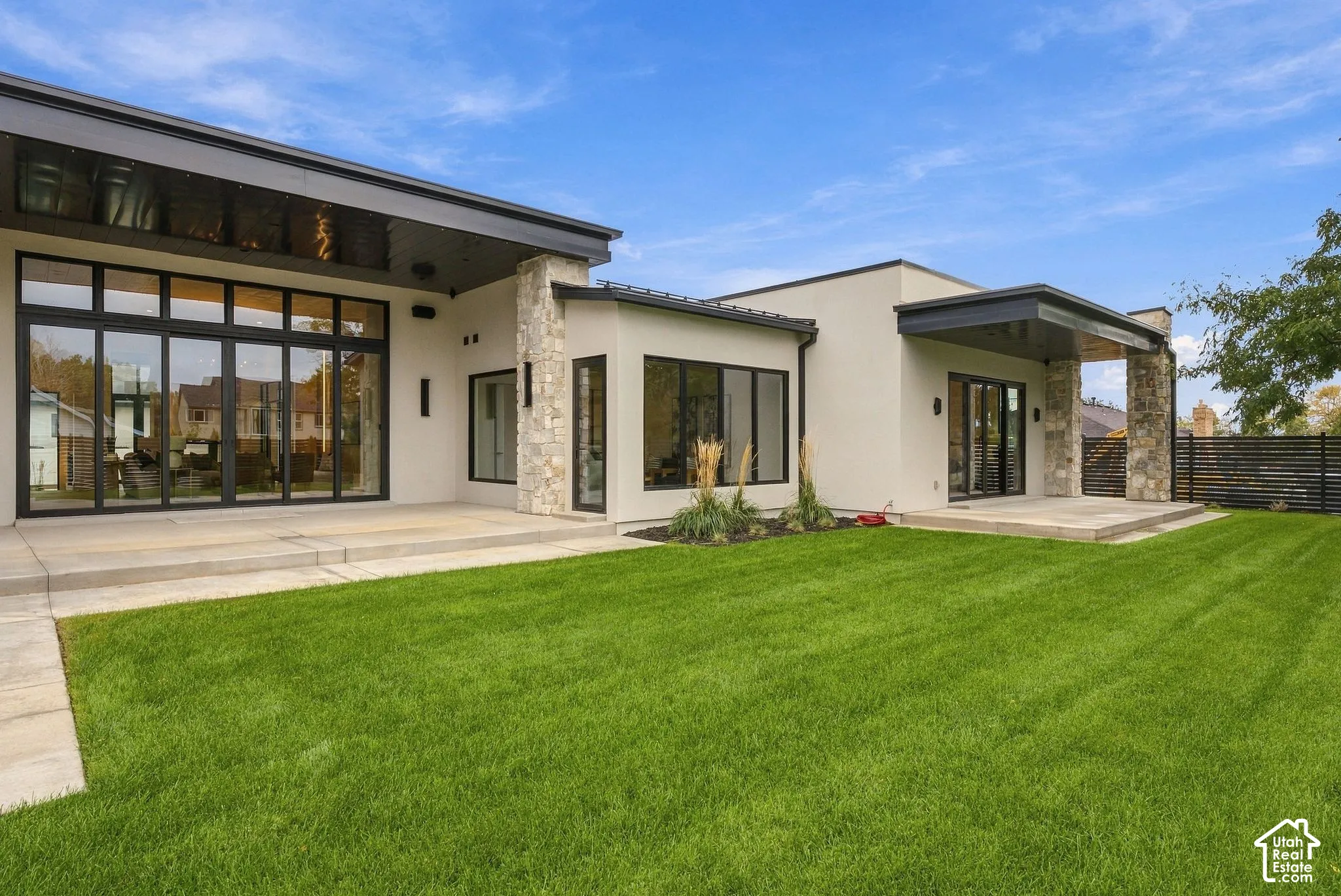 Modern exterior featuring stacked stone accents, floor-to-ceiling glass wall systems, dual covered concrete patios, and a manicured lawn