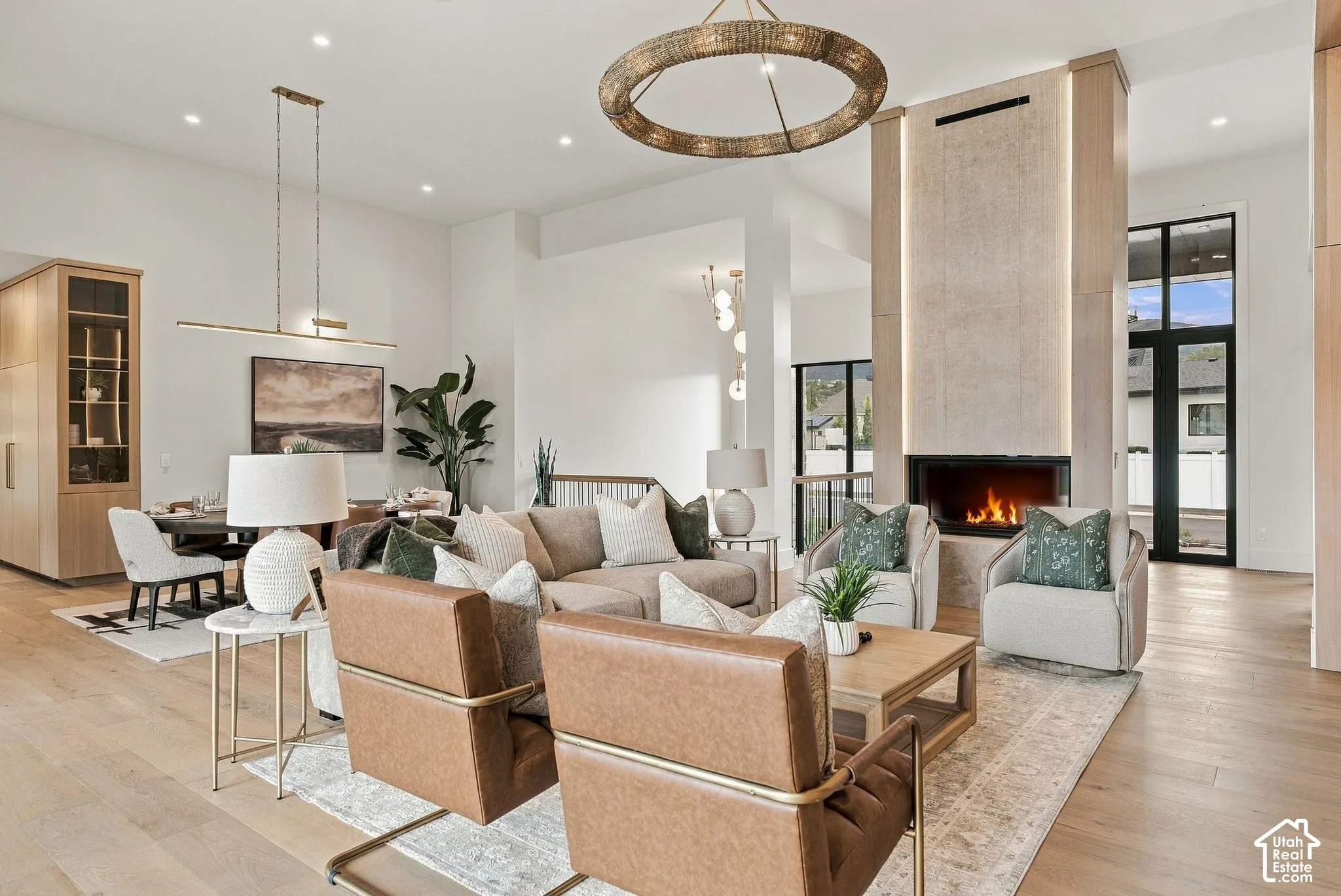 Open-concept living area featuring wood-finish flooring, a full-height white oak and stone double sided fireplace with integrated vertical lighting, a large circular statement chandelier, and floor-to-ceiling windows