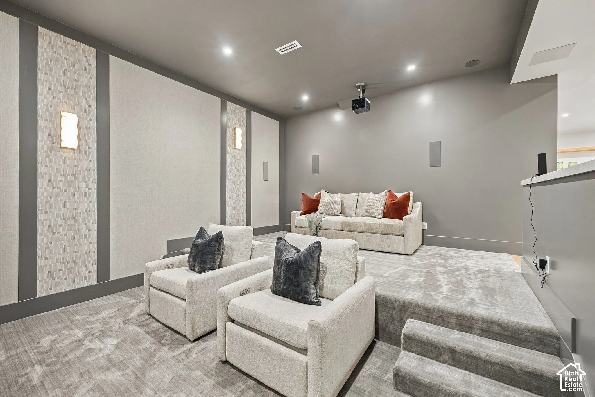 Tiered seating area with carpeted flooring, decorative wall paneling with mosaic tile inlays, built-in wall-mounted lighting, recessed ceiling lighting, and a ceiling-mounted projector