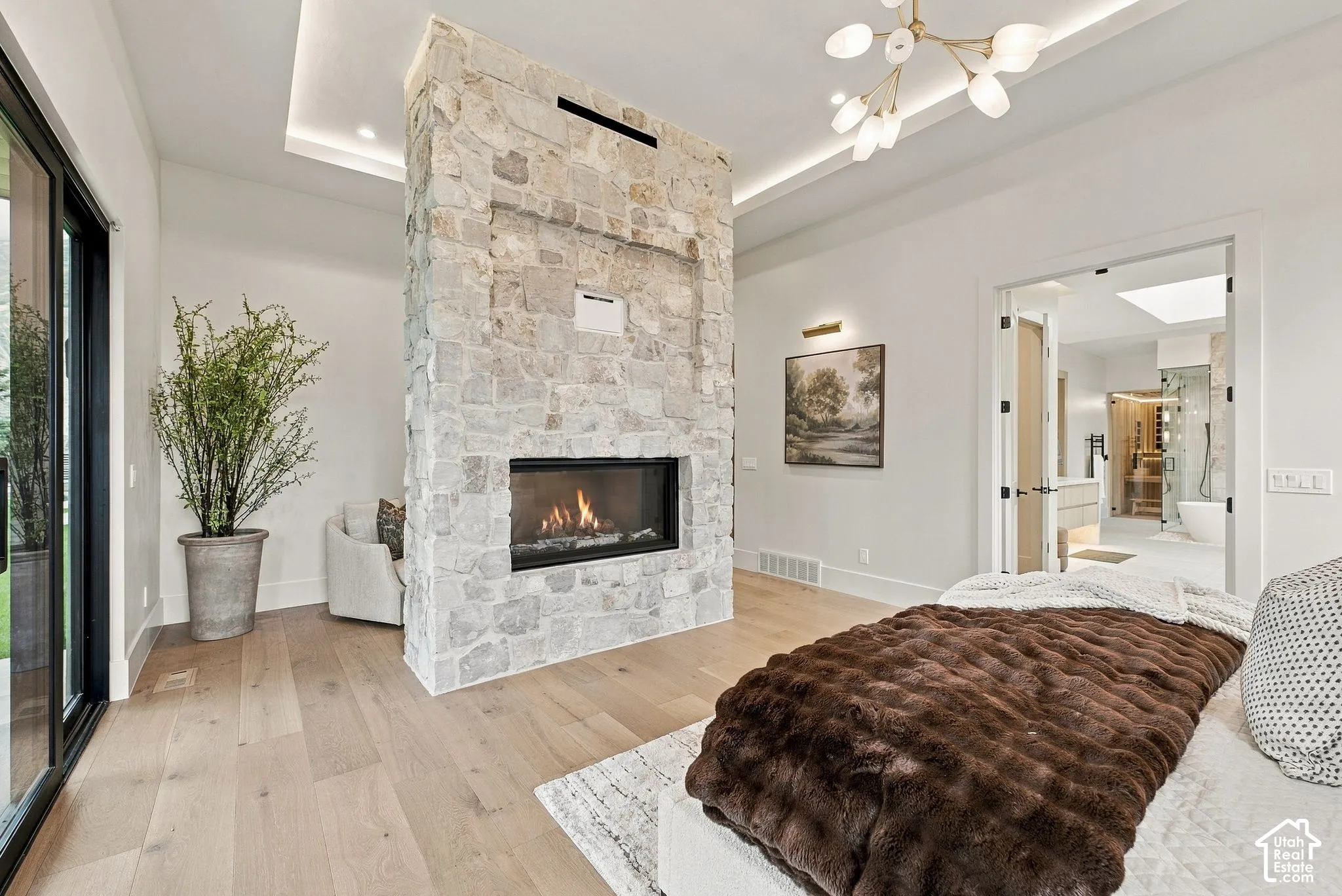 Floor-to-ceiling stone-clad double-sided fireplace, wood-finish flooring, recessed tray ceiling with integrated lighting, oversized sliding glass door, and multi-arm chandelier