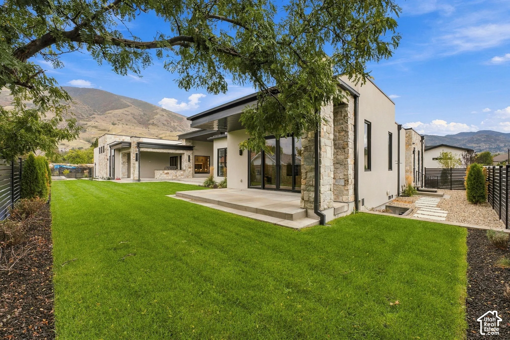 Modern stucco exterior featuring stone masonry accents, a covered concrete patio, multi-panel glass doors, and a manicured lawn set against a mountain backdrop