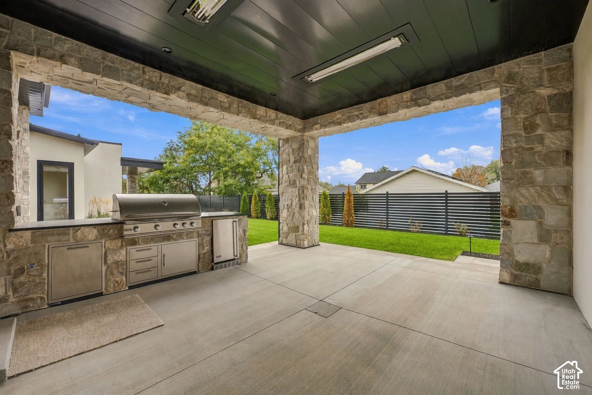 Covered outdoor patio featuring natural stone pillars, stainless steel built-in grill, ceiling-mounted heaters, and dark wood-finished ceiling panels