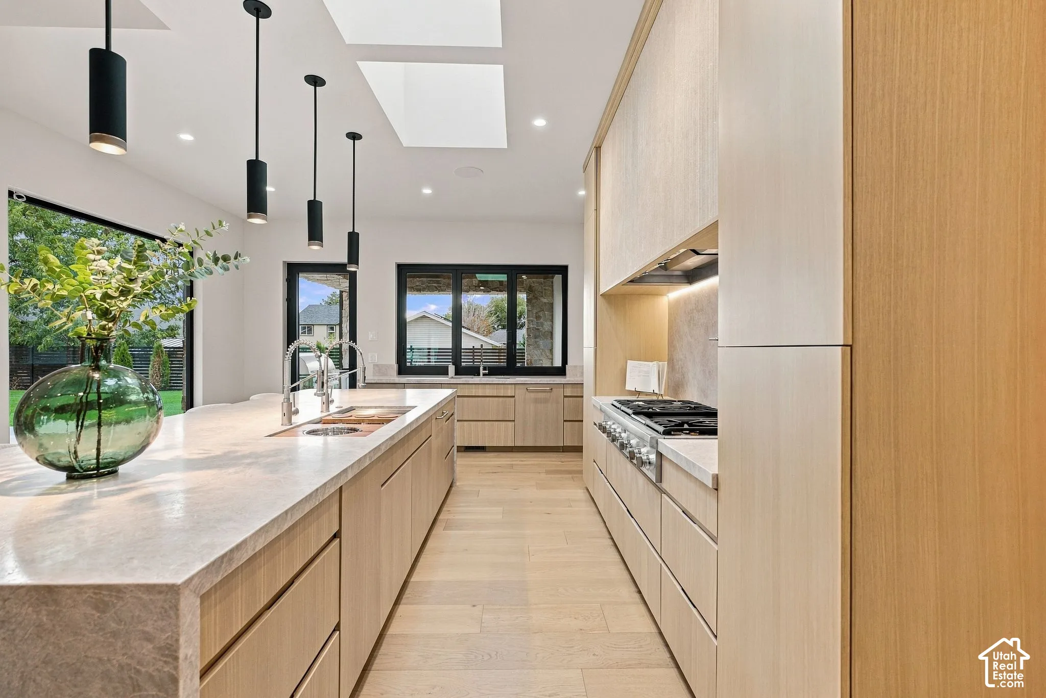 Kitchen featuring light wood-finish cabinetry, waterfall-edge stone island with an integrated sink, gas cooktop, recessed lighting, and skylights