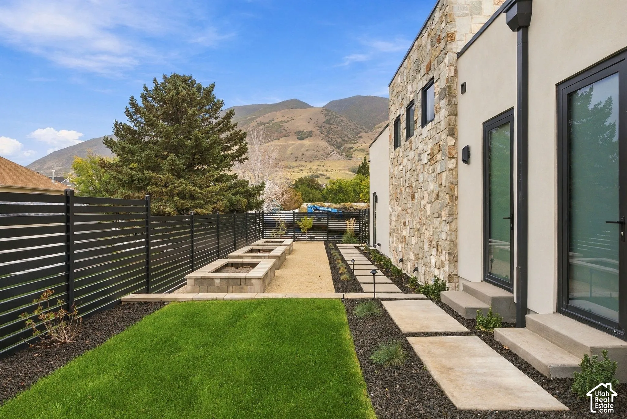 Exterior featuring a natural stone facade, horizontal metal privacy fencing, tiered concrete planter boxes, manicured lawn, and concrete paver walkway