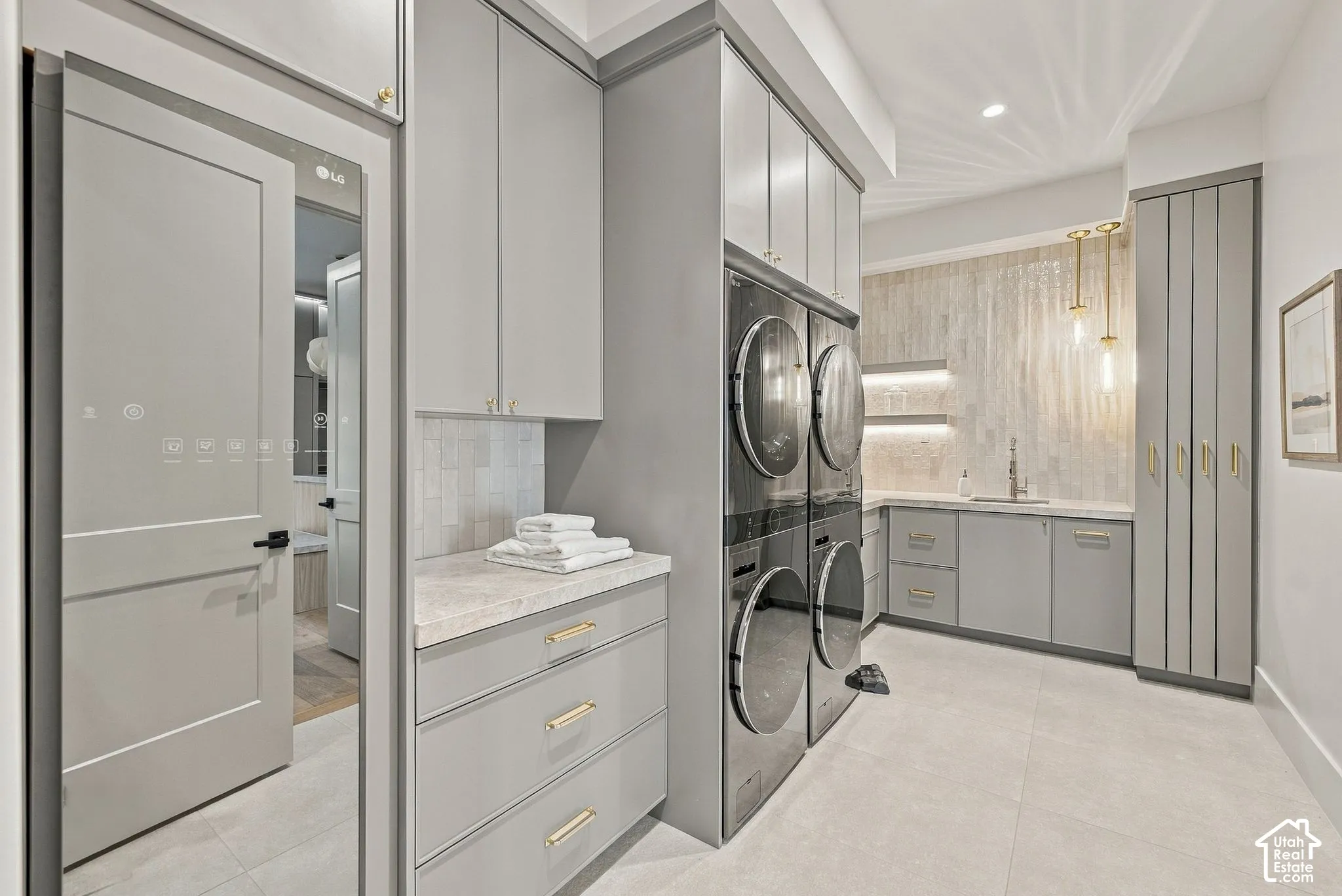 Utility area featuring custom floor-to-ceiling cabinetry, light-toned stone countertops, built-in washer and dryer set, undermount sink with brass hardware, and floor-to-ceiling vertical tile backsplash