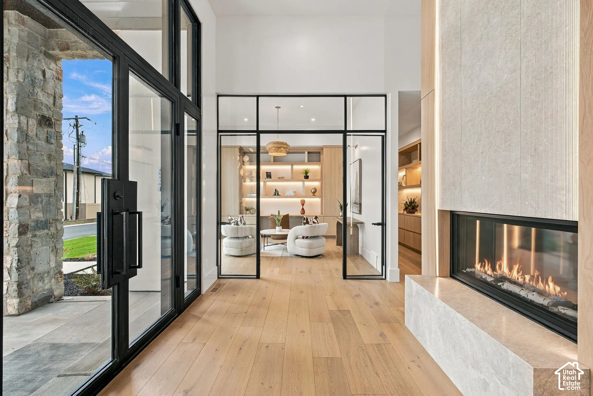 Entryway featuring natural stone exterior masonry, floor-to-ceiling glass and steel entry doors, wood-finish flooring, a linear fireplace with a stone-surround hearth, and glass-enclosed office space with built-in cabinetry