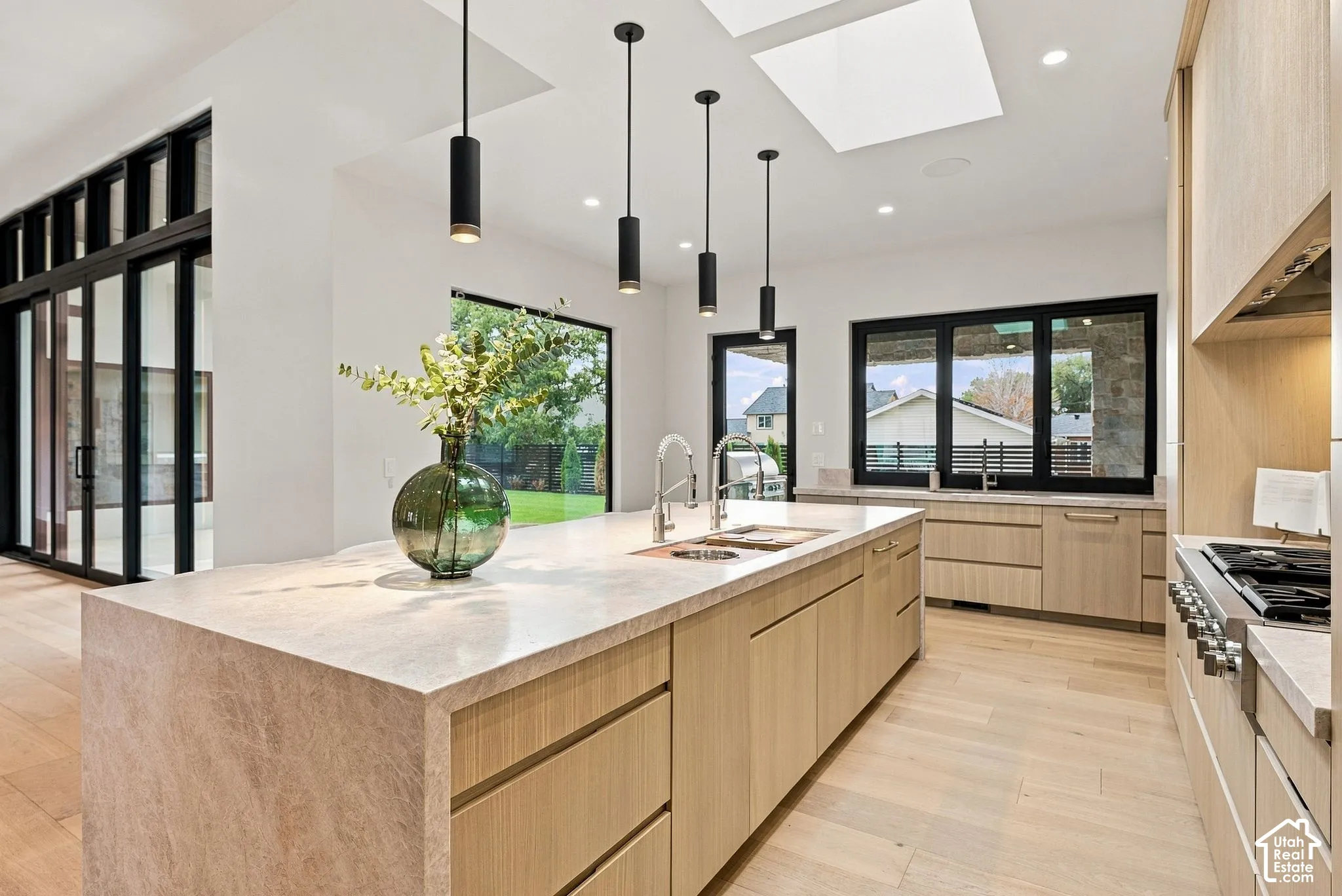 Kitchen featuring light-finish cabinetry, a large waterfall island with integrated dual sinks, sleek cylinder pendant lighting, skylights, and expansive window groupings with black framing