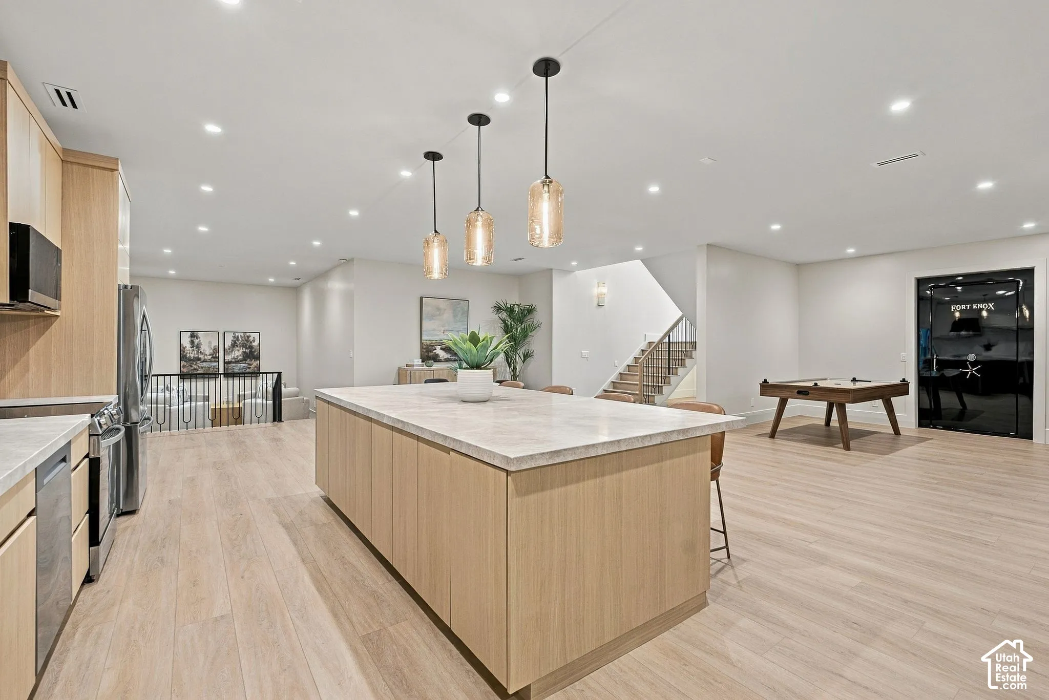 Open-concept kitchen featuring light wood-finish cabinetry, a large central island with a stone-finish countertop, recessed lighting, and three cylindrical pendant lights