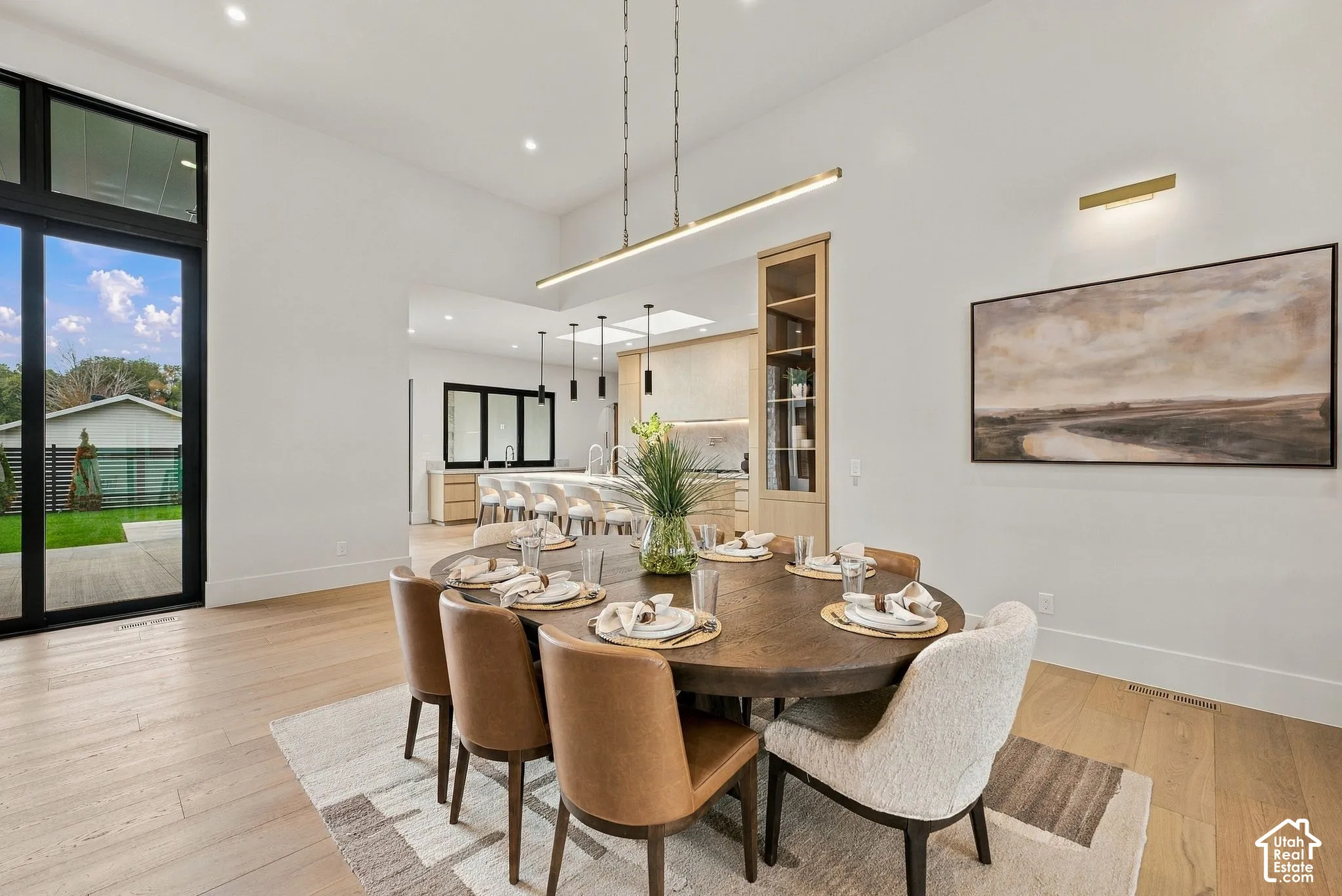 High-ceiling dining area featuring light-toned wood-finish flooring, a linear gold-tone pendant light, a built-in floor-to-ceiling display cabinet, and black-framed sliding glass doors