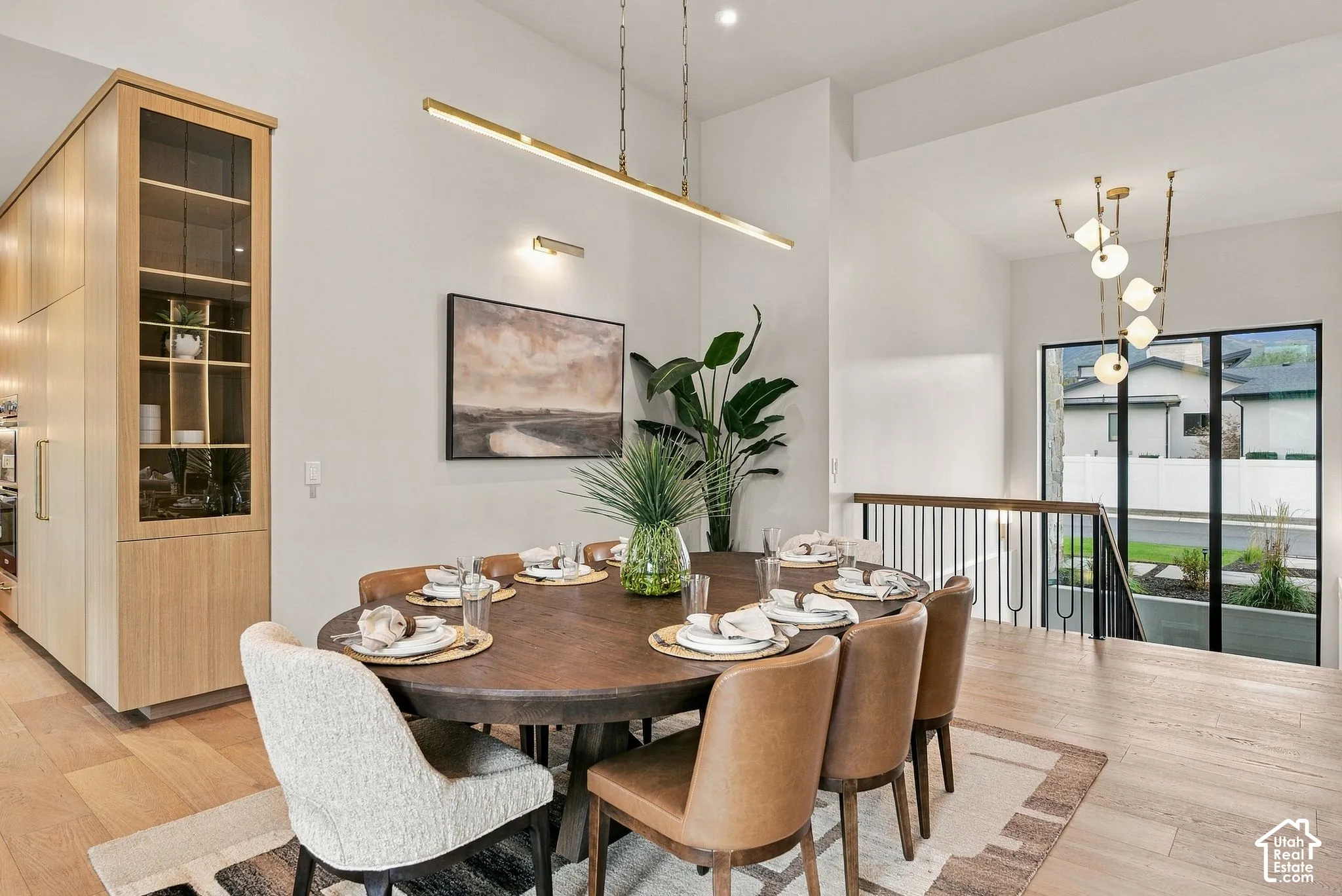Dining area featuring high ceilings, light-tone wood-finish flooring, floor-to-ceiling built-in white oak cabinetry with glass-fronted shelving, contemporary gold linear pendant lighting, and black-framed windows
