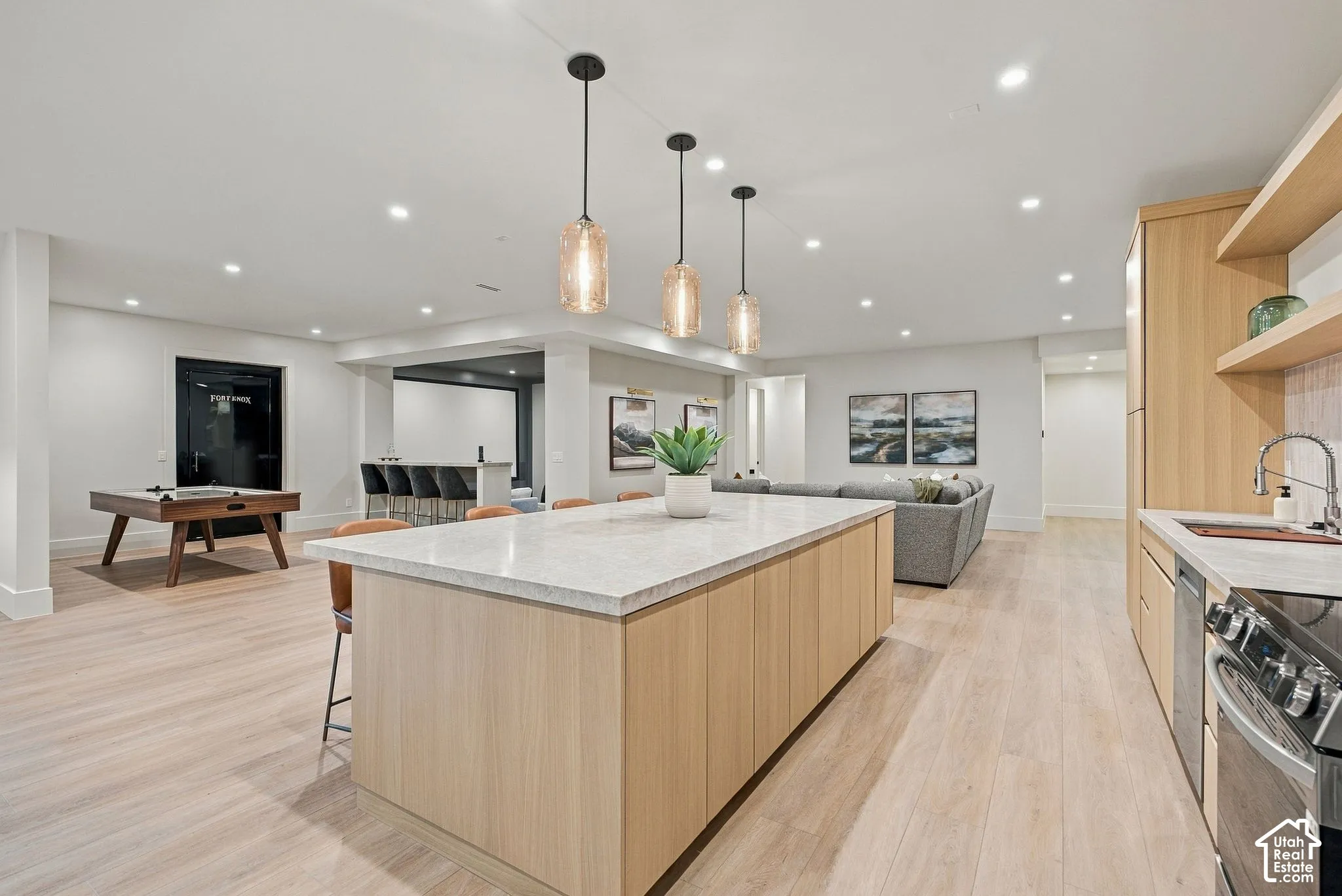 Open-concept space featuring light-toned wood-finish cabinetry, an expansive kitchen island with a stone countertop, recessed lighting, and trio of glass pendant lights
