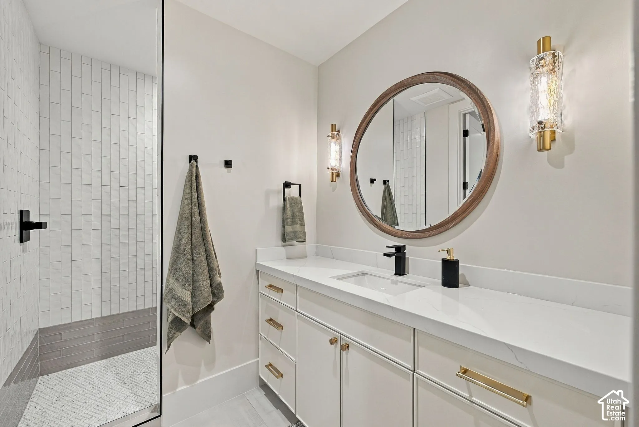Bathroom featuring a white vanity with integrated countertop, round wall-mounted mirror, vertical textured tile shower surround, matte black hardware, and tiered brass-finish wall sconces