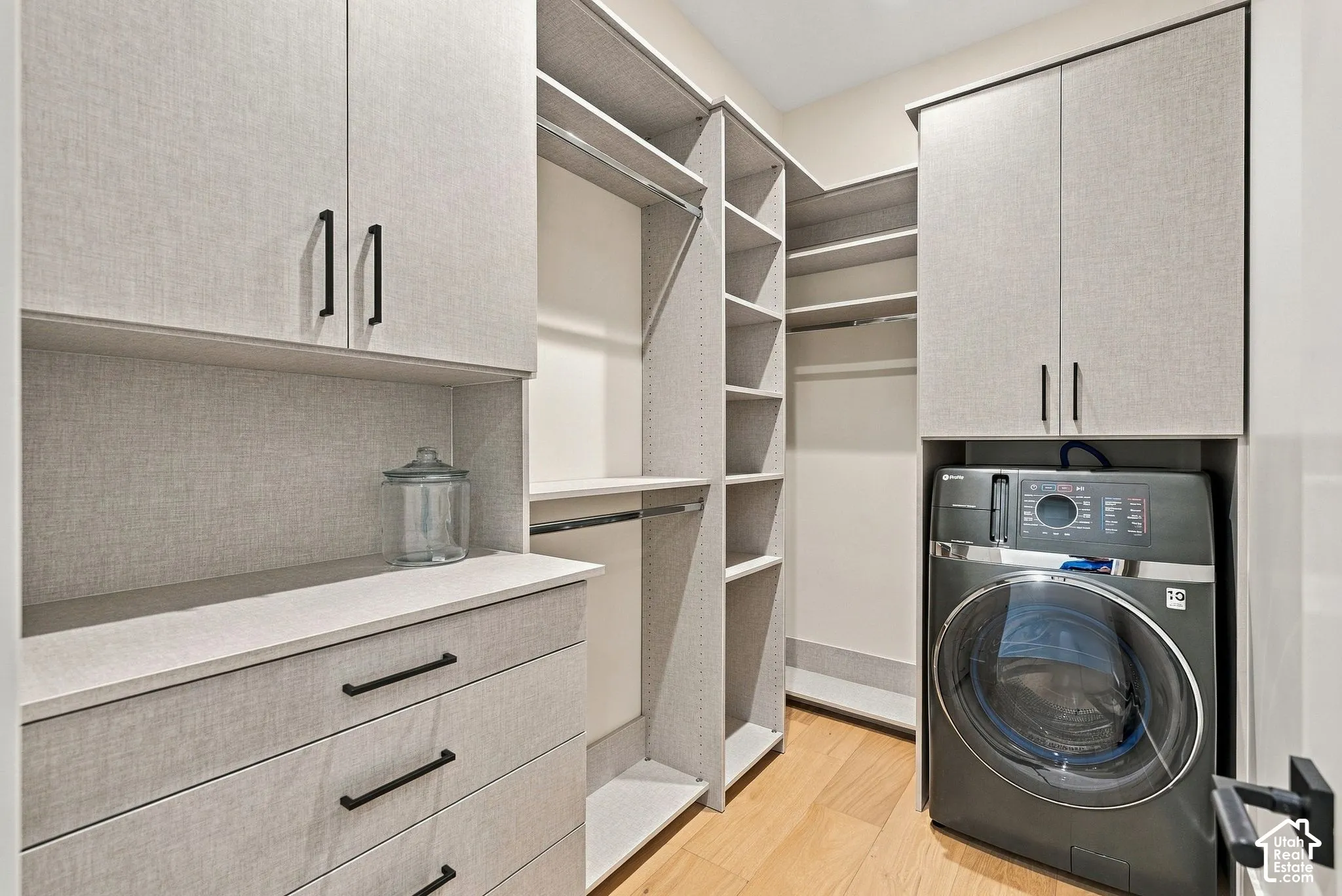 Custom storage space featuring light-toned cabinetry, built-in shelving, integrated dresser drawers with matte black hardware, and an alcove for laundry appliances over wood-finish flooring