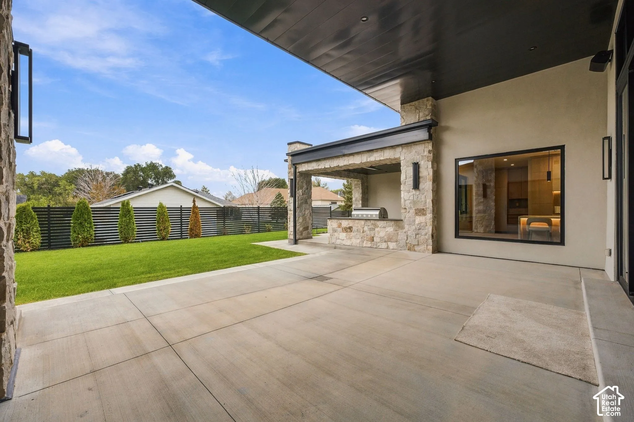 Covered patio featuring concrete flooring, stone-clad outdoor kitchen, recessed lighting, and expansive lawn with perimeter privacy fencing