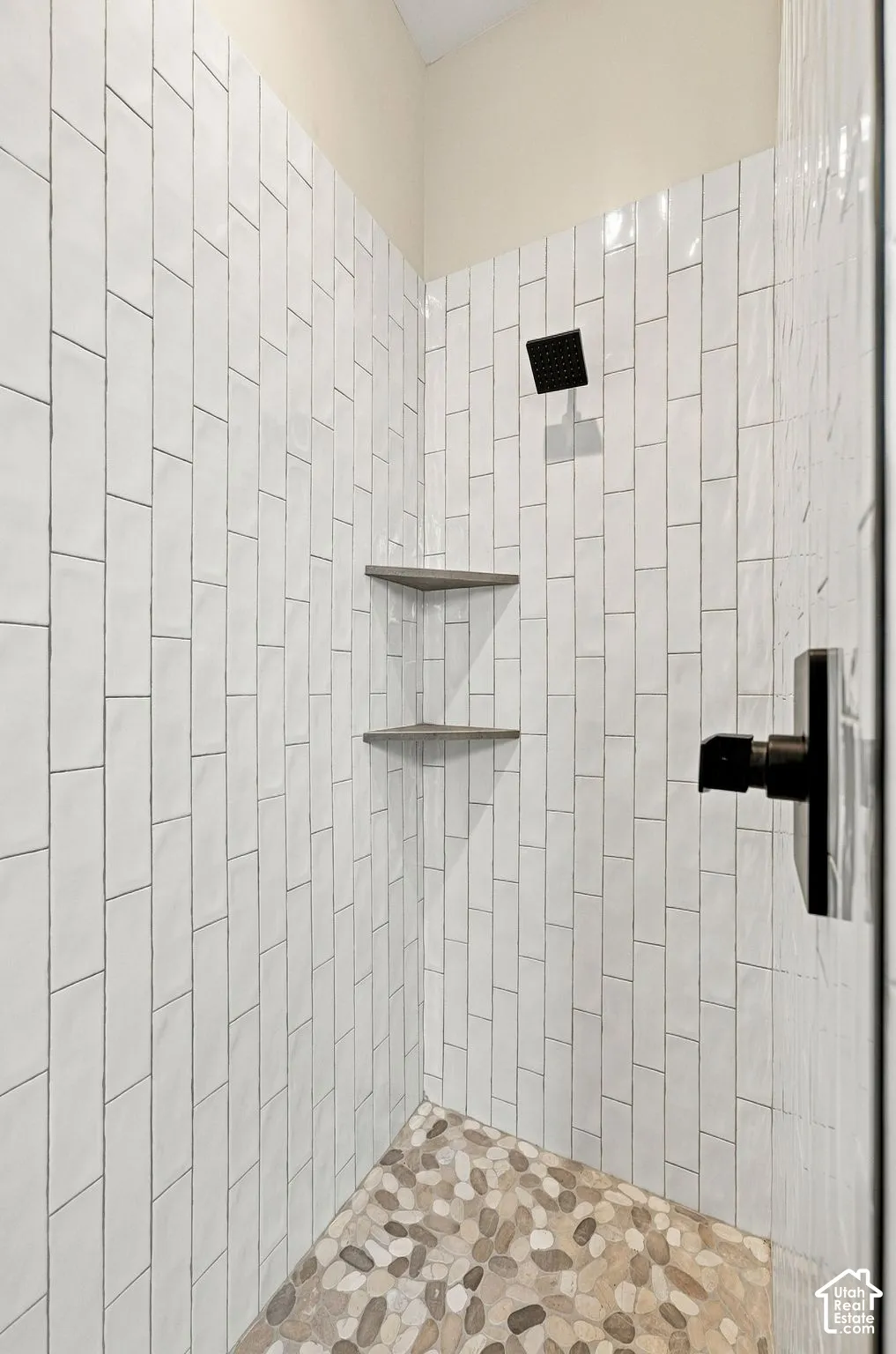 Shower enclosure featuring vertical subway tile walls, a pebble stone floor, matte black hardware, and dual built-in corner shelves