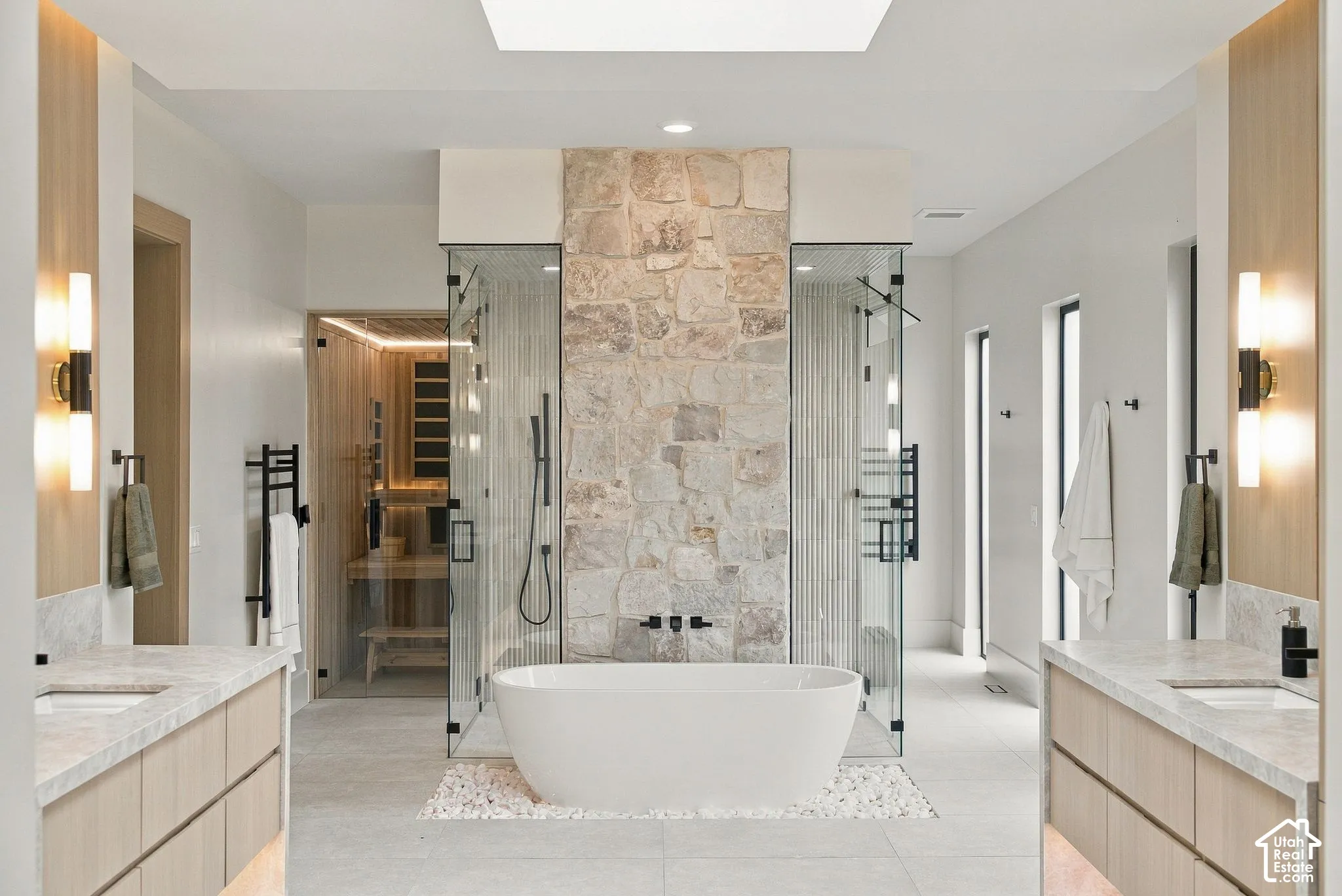 Freestanding oval soaking tub set on a stone-tile base, glass-enclosed dual showers with a central natural stone accent wall, light wood-finish cabinetry with stone countertops, and integrated wall-mounted lighting