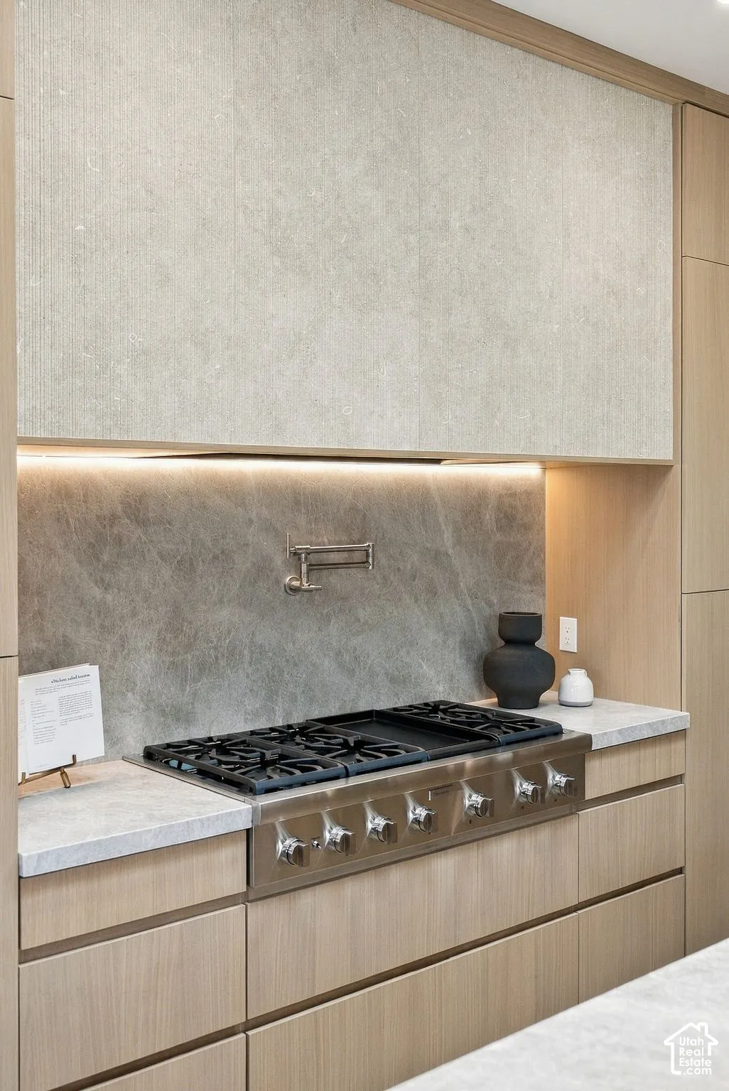 Custom wood cabinetry with flat-panel doors, professional-grade gas range, stone-slab backsplash, wall-mounted pot filler faucet, and integrated under-cabinet lighting