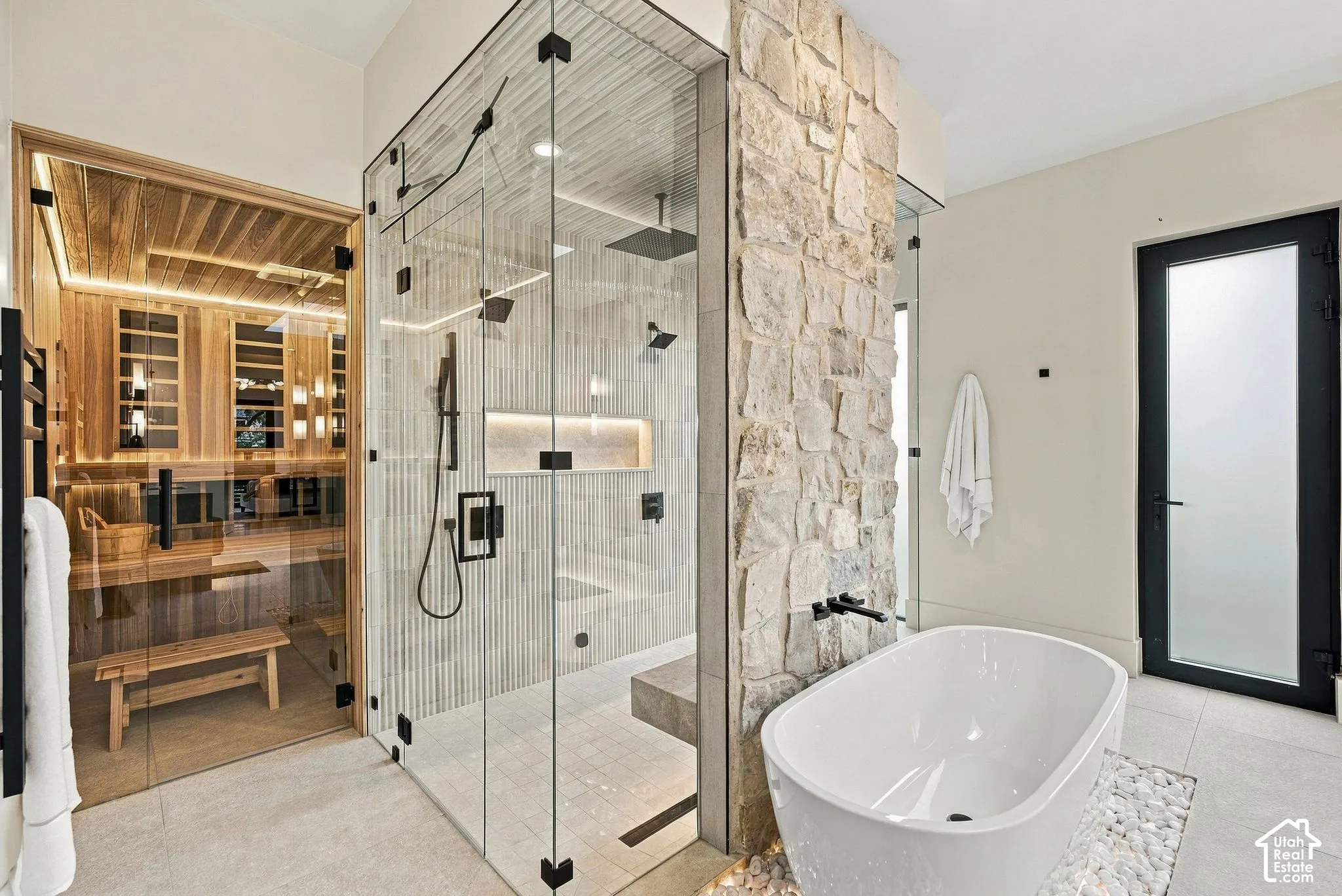 Frameless glass sauna with wood-panel interior, walk-in shower with vertical tile surround and recessed lighting, natural stone accent wall, freestanding soaking tub, and matte black plumbing fixtures