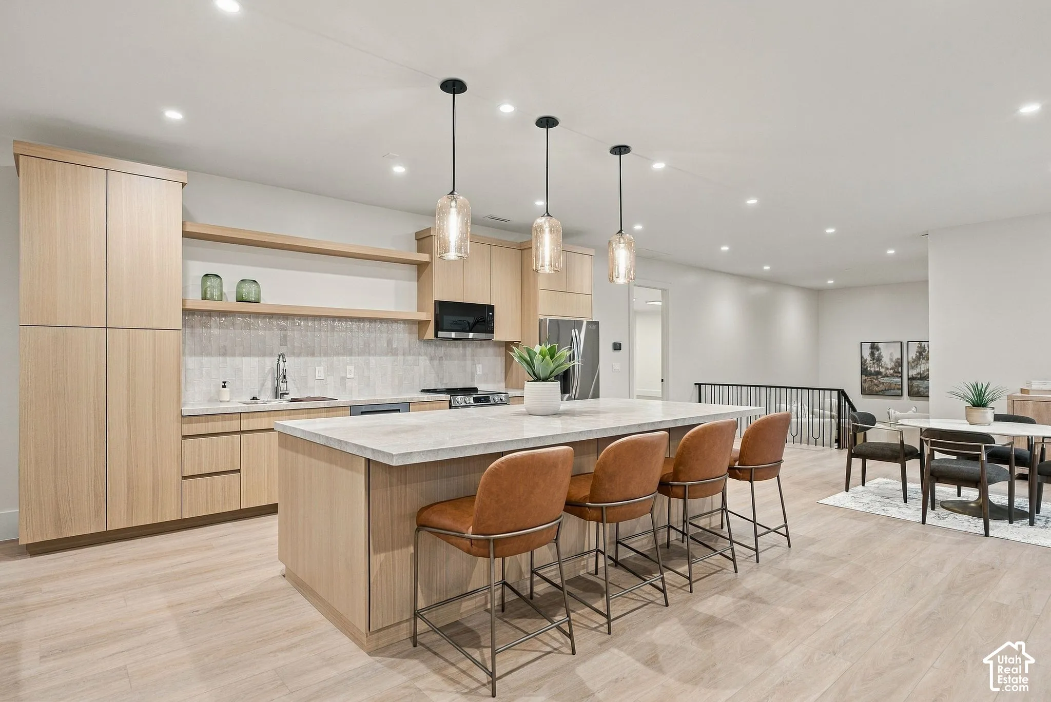 Kitchen featuring light wood-finish cabinetry, a large central island with a stone-surface countertop, textured tile backsplash, open shelving, and three glass pendant lights