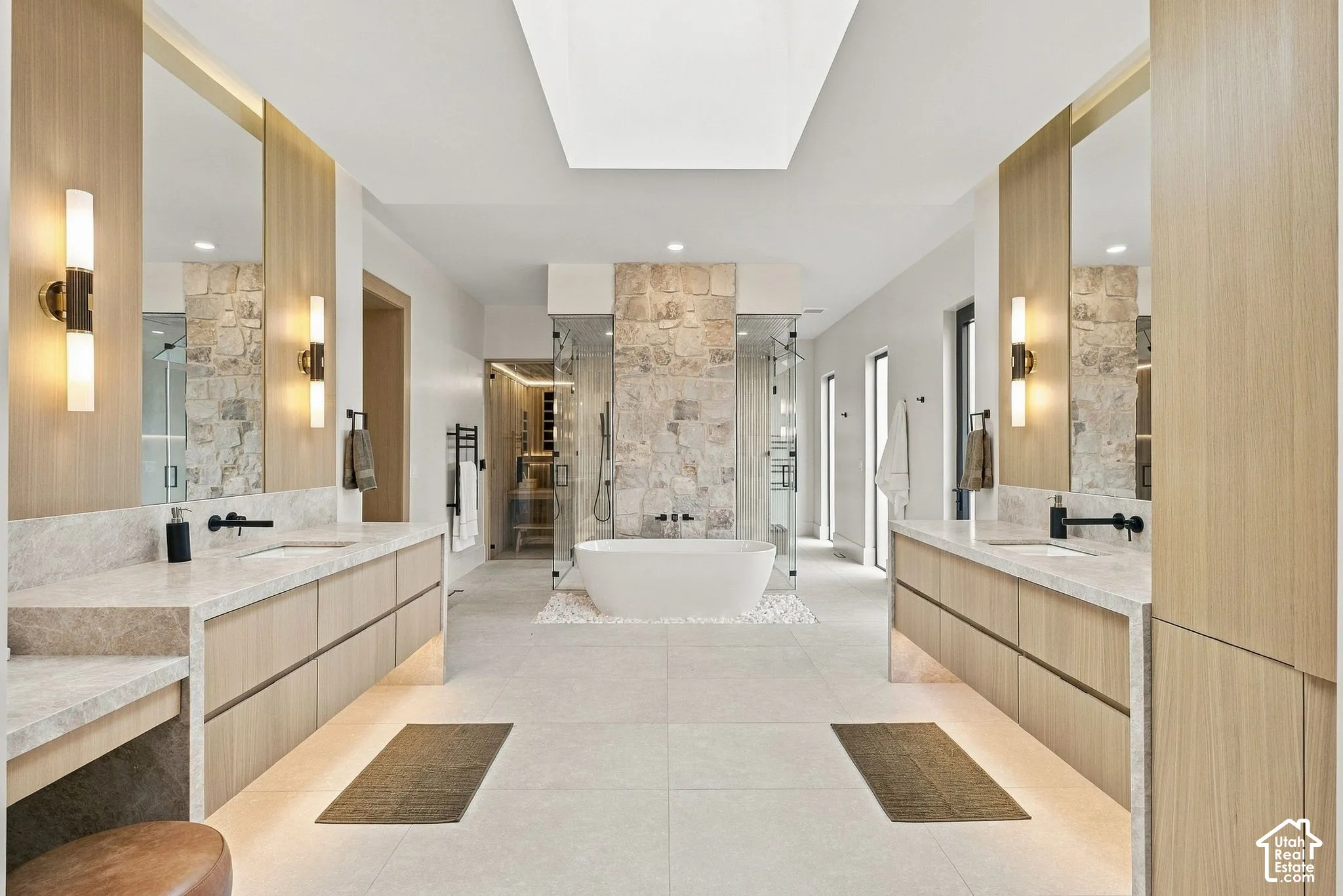 Dual vanities with light wood cabinetry, stone countertops, integrated lighting, wall-mounted matte black faucets, and a freestanding soaking tub centered against a stone-accented wall beneath a skylight