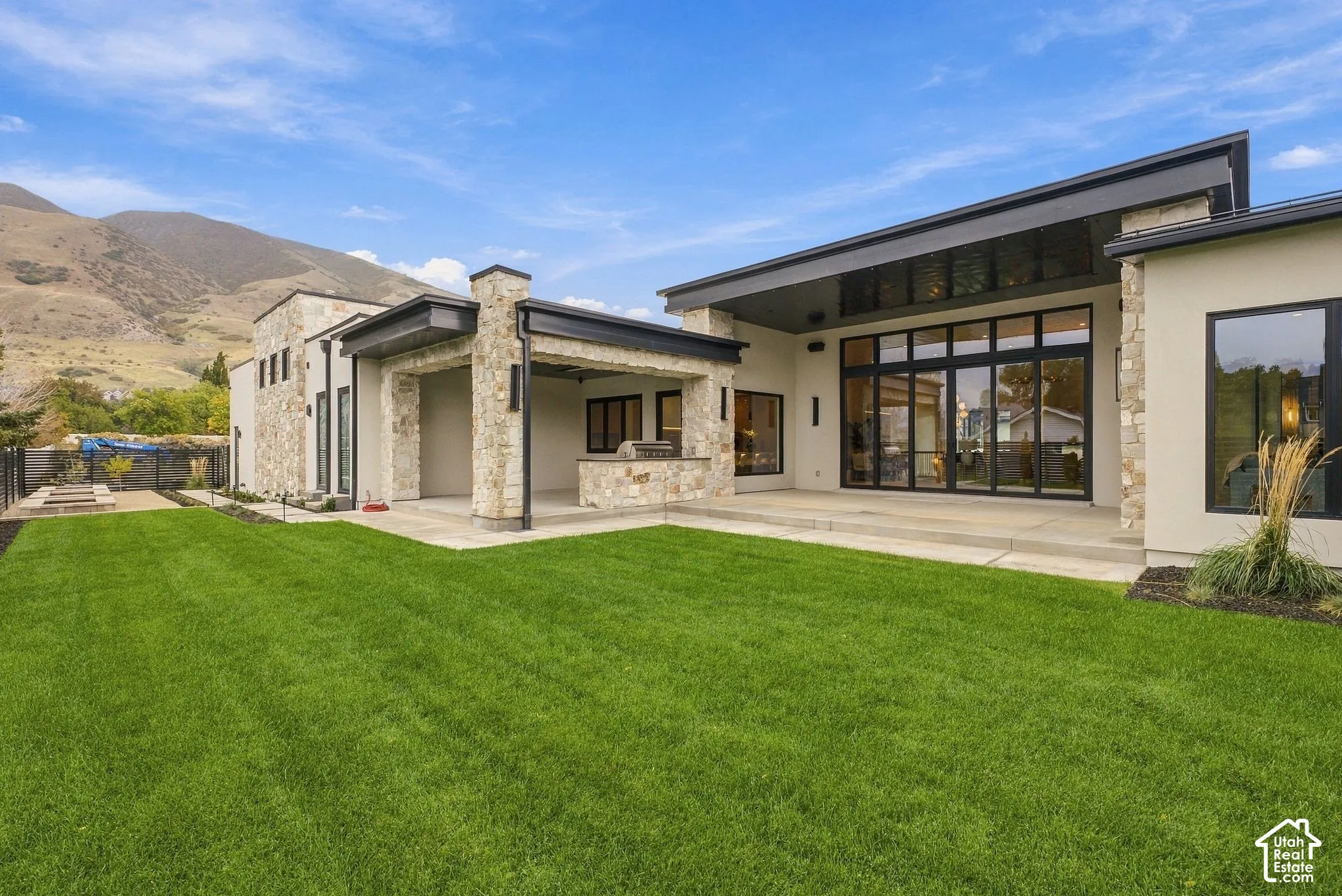 Modern residence exterior featuring stone masonry, expansive glass wall panels, a covered outdoor kitchen, and concrete patio flooring set against a mountain backdrop
