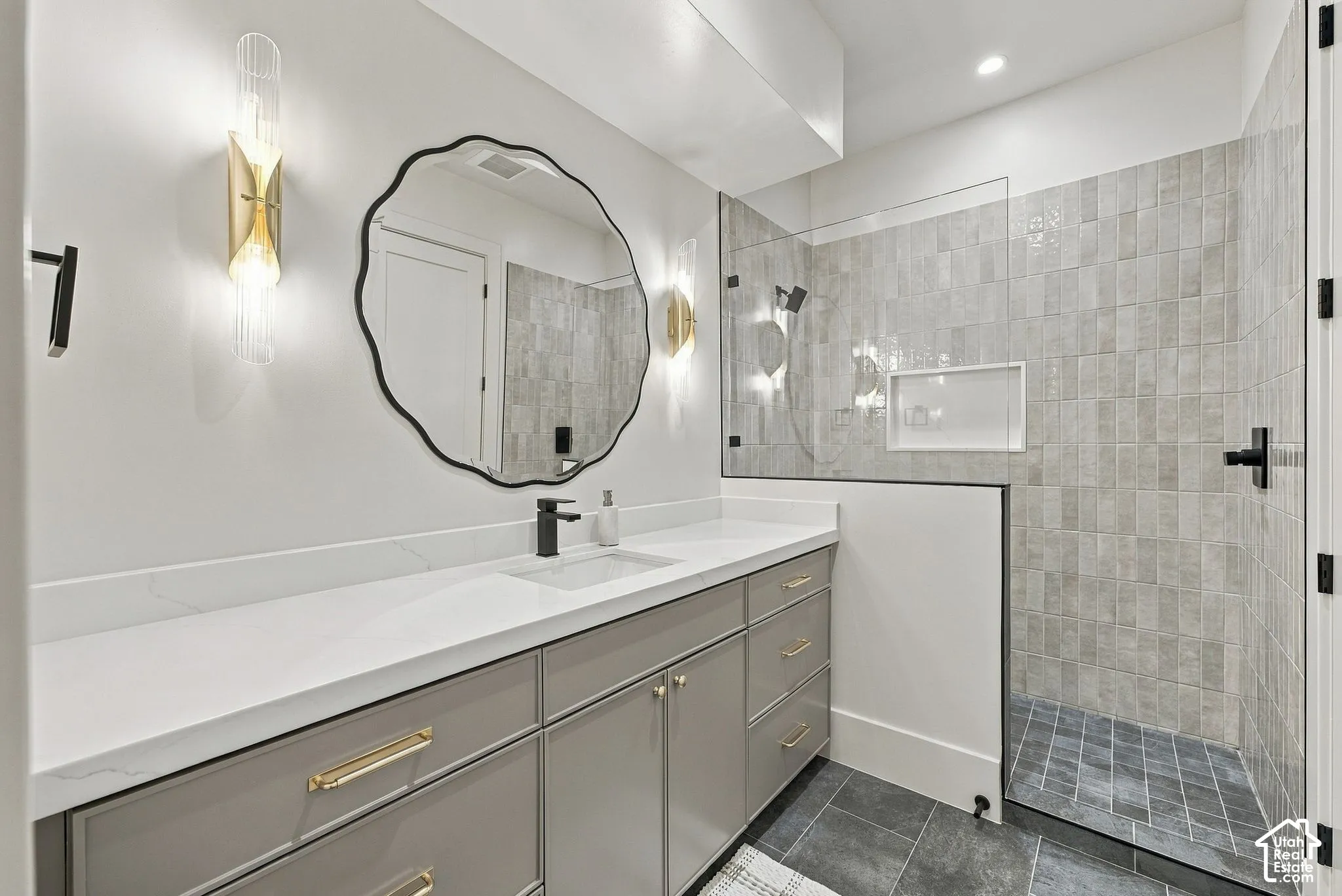 Bathroom featuring a vanity with quartz countertop, dual gold-finish wall sconces, a custom-shaped mirror, matte black fixtures, and a walk-in shower with vertical textured tile and built-in recessed shelving