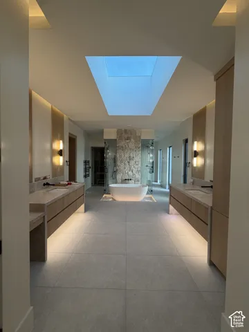 Spacious spa inspired primary  bathroom featuring a central skylight that floods the room with natural light,  freestanding soaking tub, stone accent wall, and dual floating vanities with integrated sinks and sconce lighting, heated floors, his and hers t
