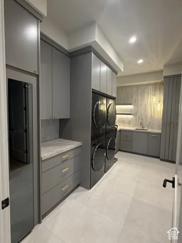 Utility room with dual access from the primary closet and the hallway featuring extensive built-in cabinetry, double stacked washer and dryer units, a workstation with a sink, and light-toned tile flooring. Features a full size clothing steamer and built