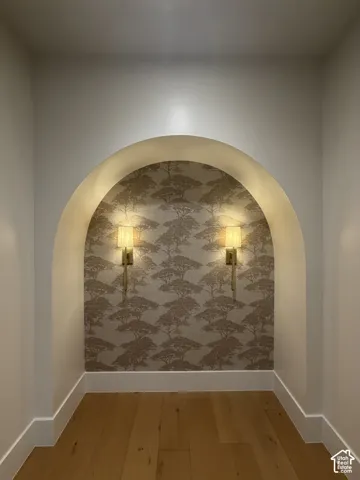 Entrance to primary bedroom, Arched wall niche featuring decorative wallpaper, two wall-mounted sconces, and wood-finish flooring with white baseboards