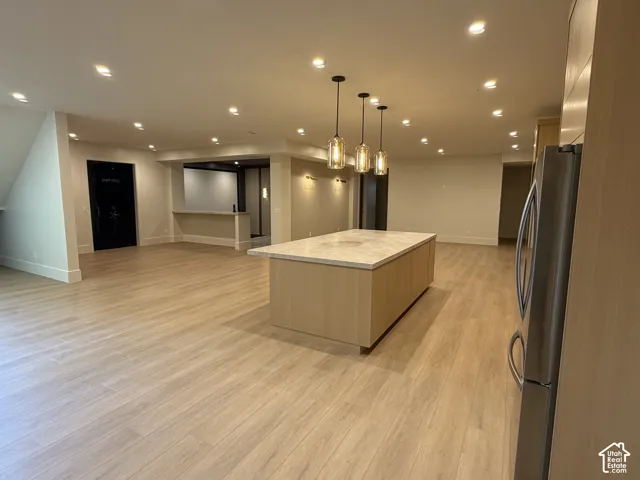 Spacious open-concept area featuring a large kitchen island with a light-toned countertop, three pendant lights, recessed lighting, wood-finish flooring, and a stainless steel refrigerator