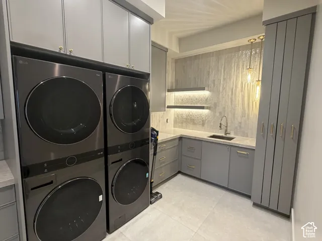 Laundry area featuring stacked appliances, grey cabinetry with gold-tone hardware, a sink with a stainless steel faucet, and a stone-finish backsplash