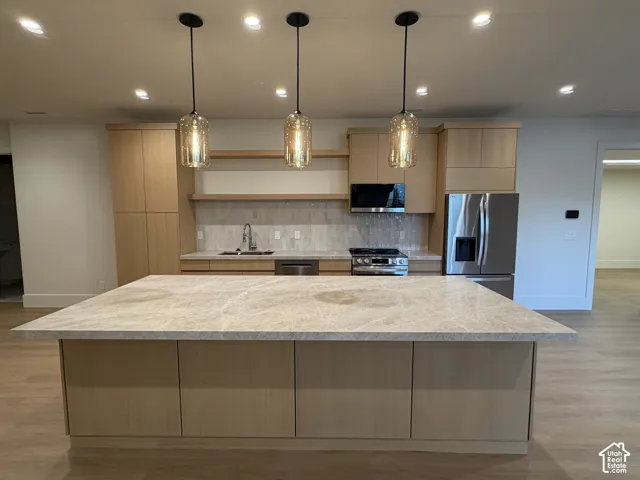 Spacious kitchen featuring a large central island with a stone countertop, light wood-finish cabinetry, stainless steel appliances, and pendant lighting