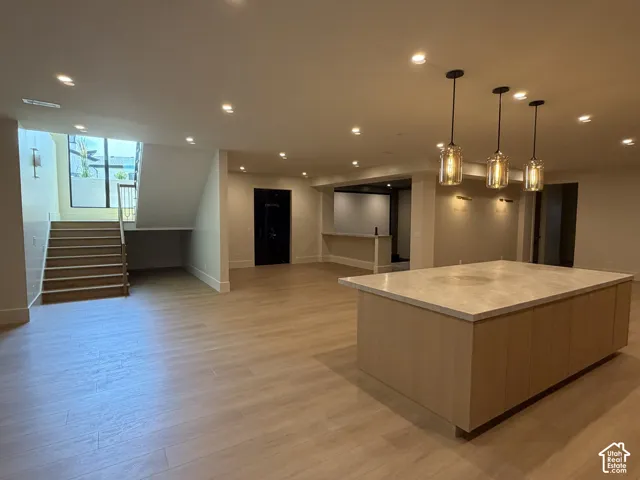 Spacious open-concept area featuring a large kitchen island with a light-toned countertop, wood-finish flooring, recessed lighting, and a prominent staircase with a window