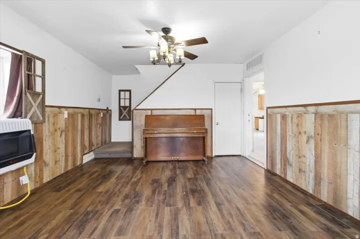 Unfurnished living room with a wainscoted wall, wood walls, heating unit, dark wood-style flooring, and a ceiling fan