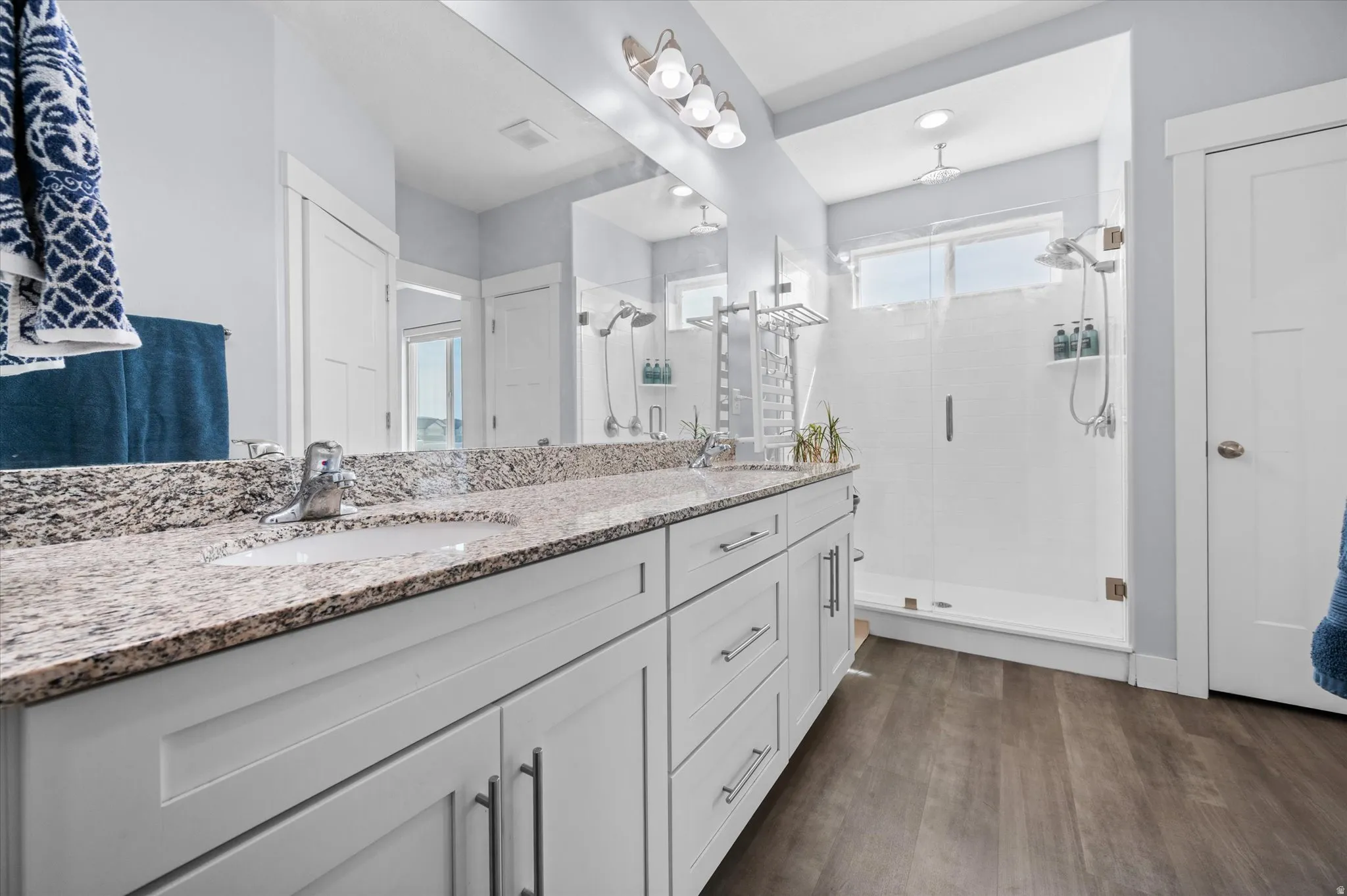 Bathroom featuring double vanity, a stall shower, with rain shower, linen closet, and dark wood finished floors