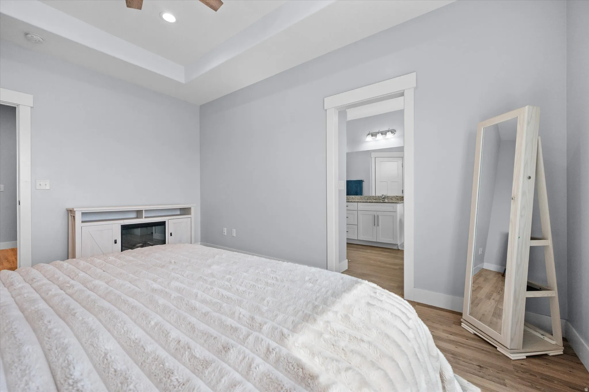 Bedroom featuring light wood-style flooring, a tray ceiling, recessed lighting, a ceiling fan, and ensuite bath