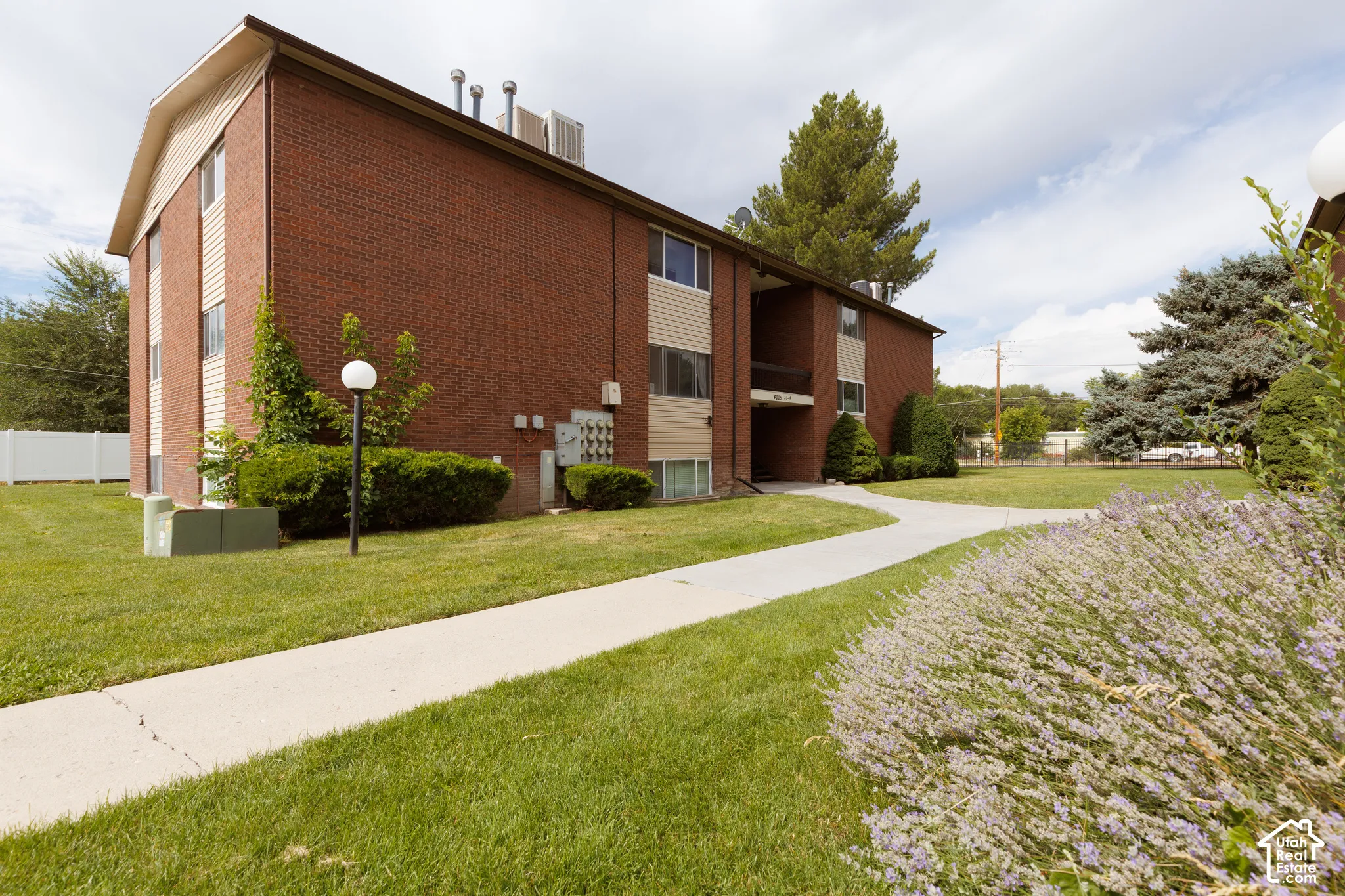 Brick exterior building with tan siding accents, featuring a concrete pathway, manicured lawn, established shrubs, and mature trees