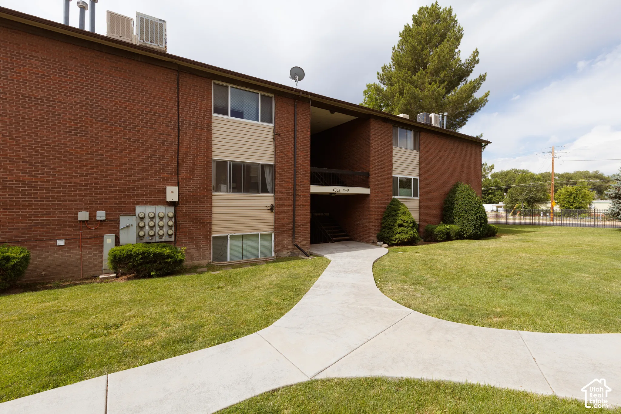 Brick-clad exterior building featuring tan siding accents, a concrete pathway, a verdant lawn, and established shrubbery
