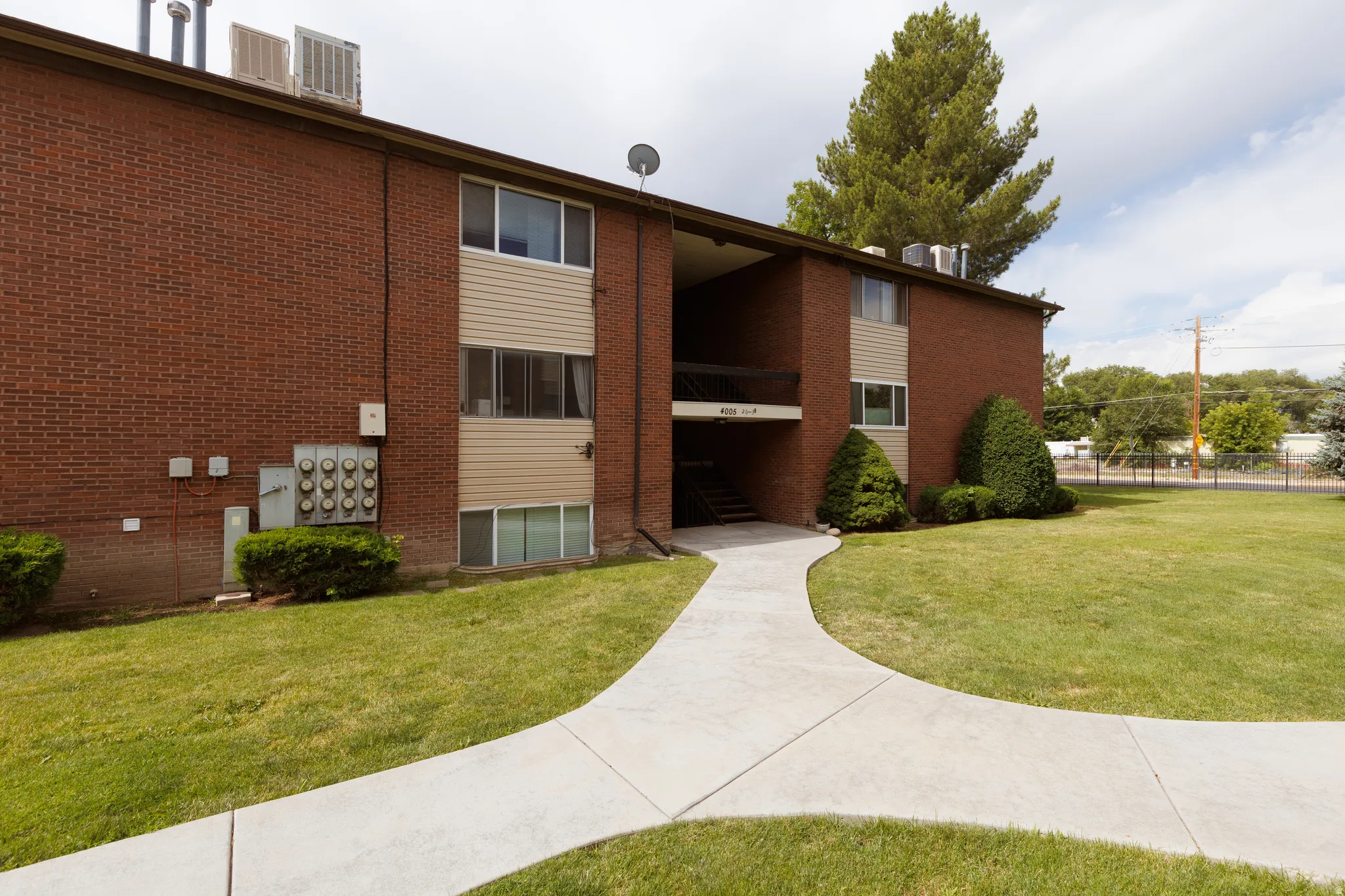 Brick-clad exterior building featuring tan siding accents, a concrete pathway, a verdant lawn, and established shrubbery